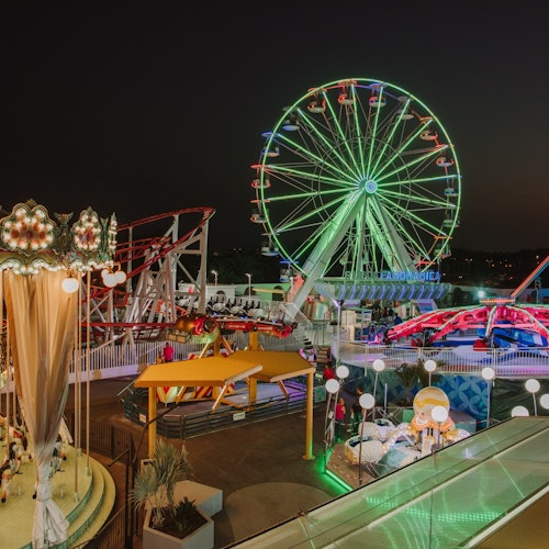 Pretpark 's nachts met verlichte reuzenrad, draaimolen, achtbanen en kleurrijke lampjes.