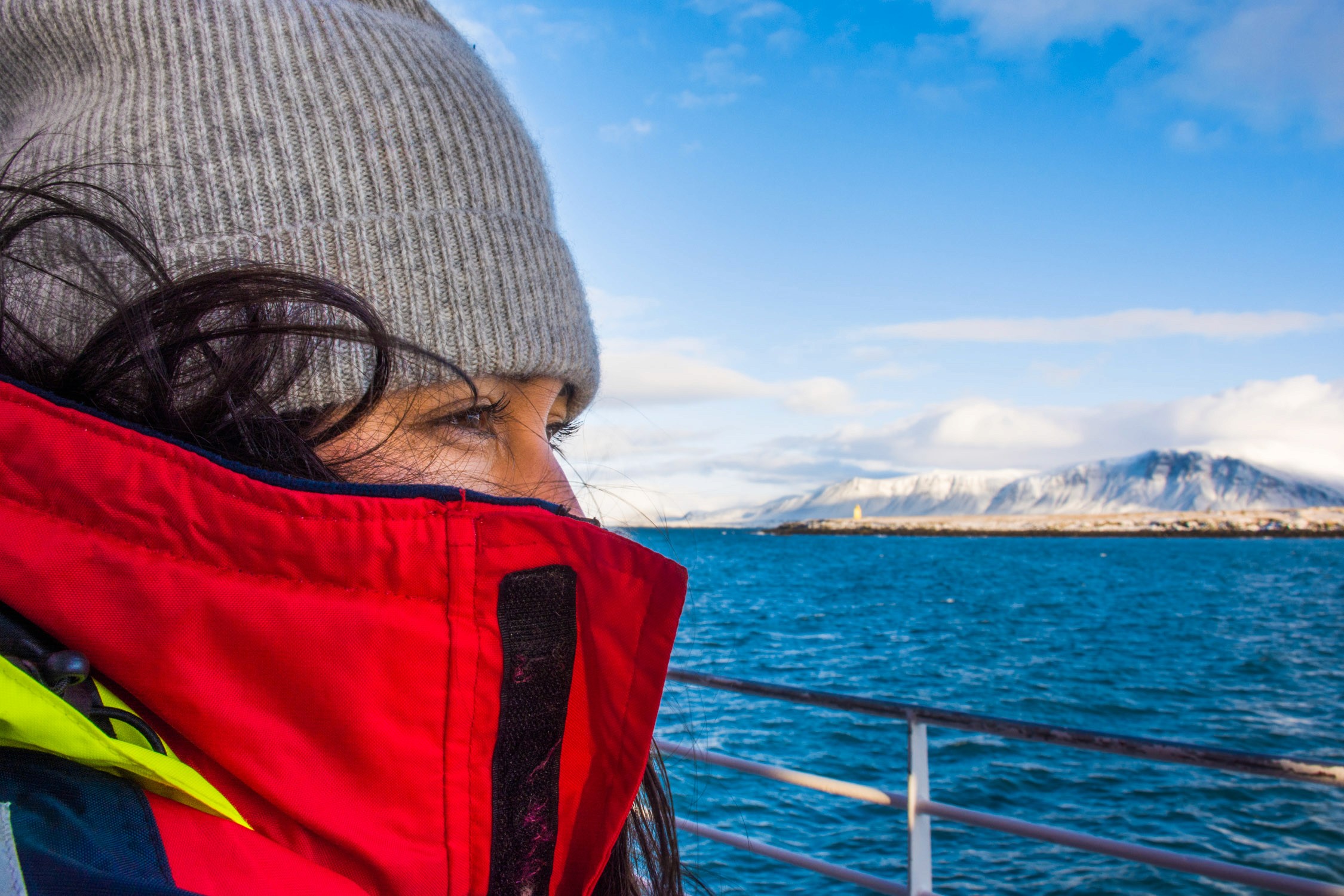 A person in a grey knit beanie and red parka looks out over a blue sea toward snow-capped mountains under a clear sky.