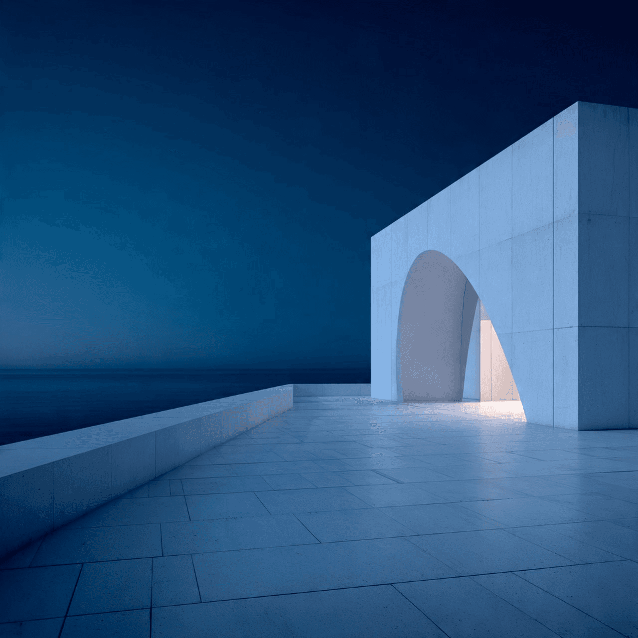 Modern minimalist building by the sea at dusk, with sleek white walls, glowing interior featuring a red chair, and a calm, serene atmosphere.