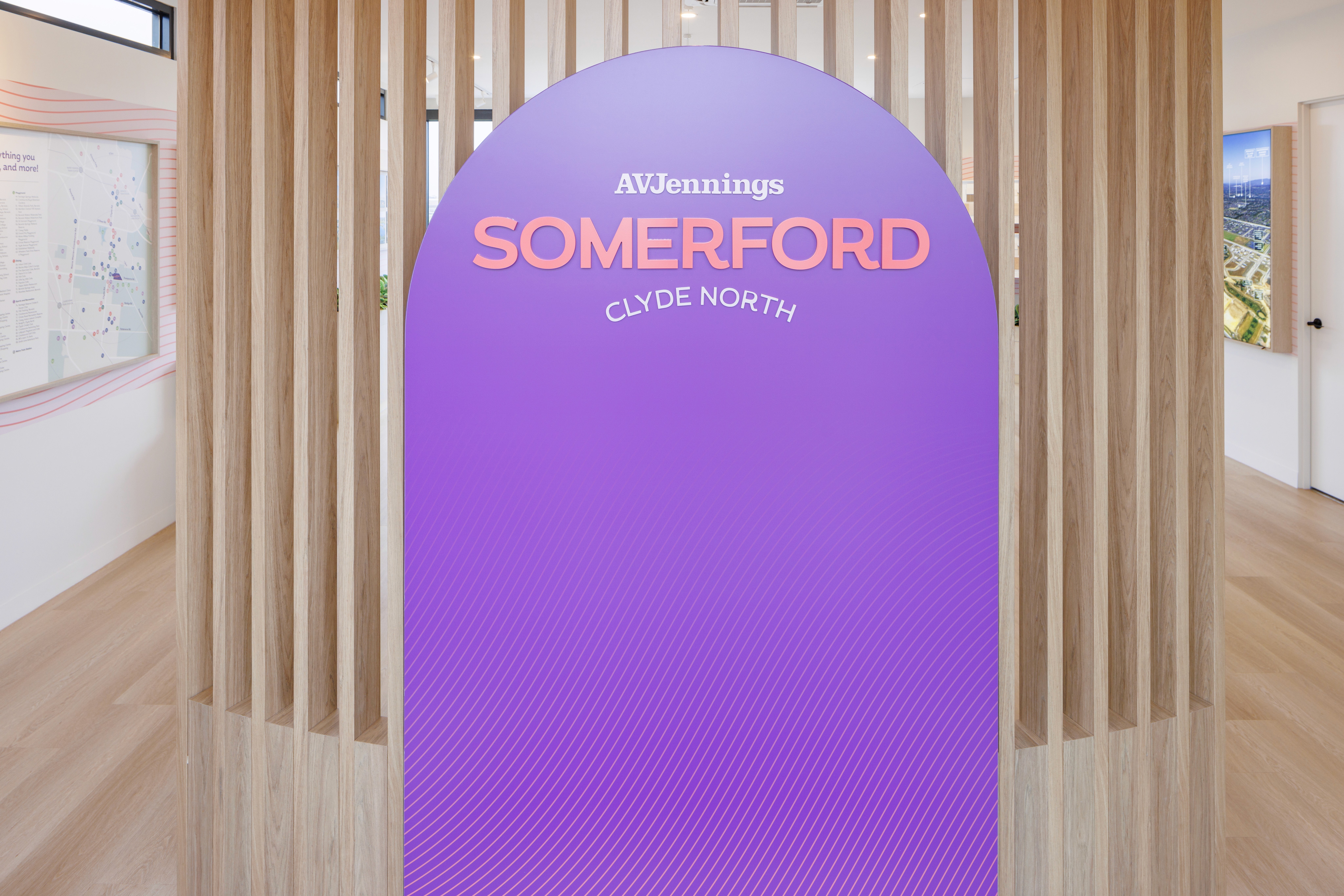 Purple sign with white and orange text reading "AVJennings, Somerford, Clyde North" in a modern room with light wood paneling and maps.