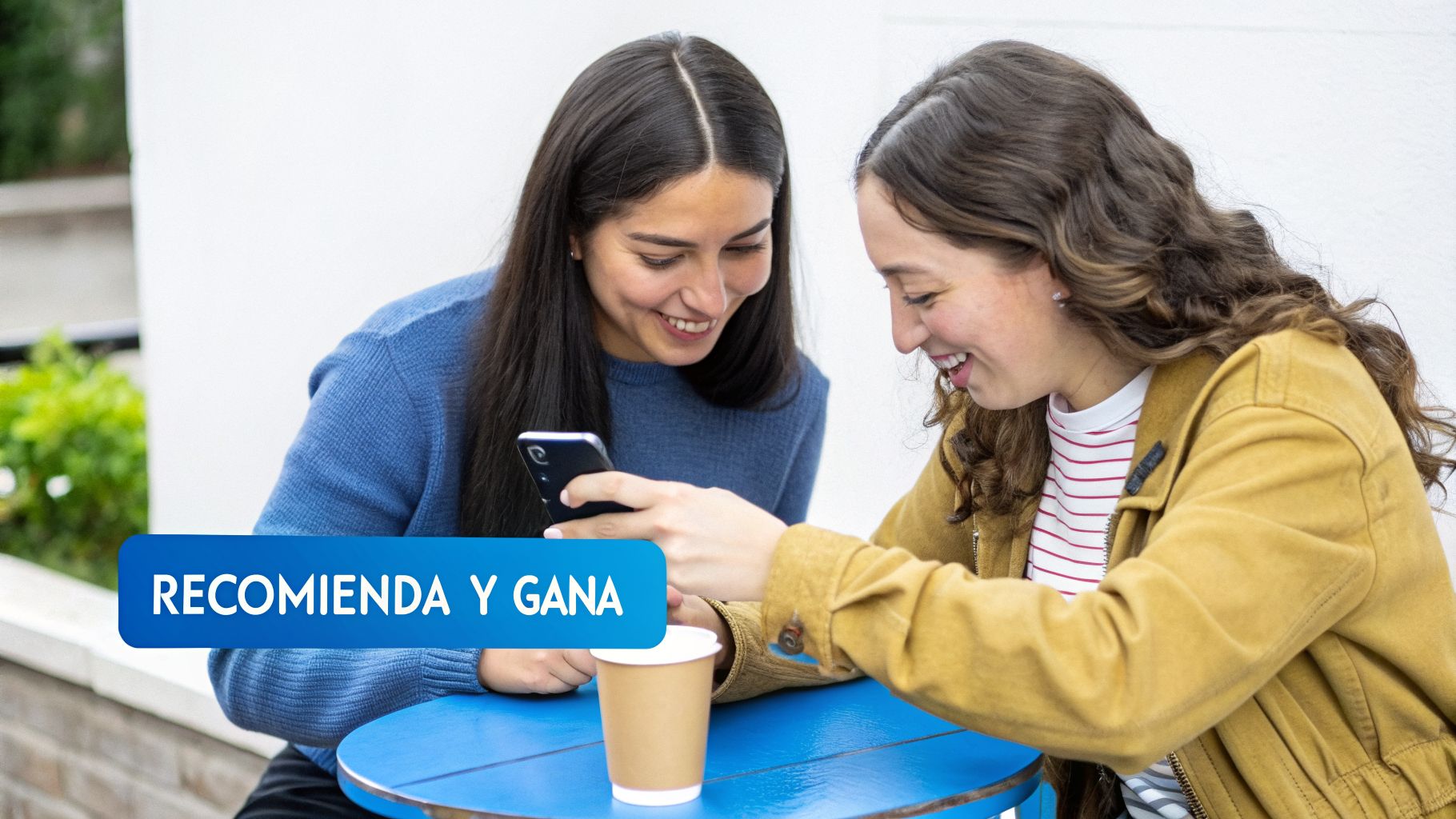 Dos mujeres sonrientes comparten un momento mirando un teléfono móvil, sentadas en una mesa azul con café.