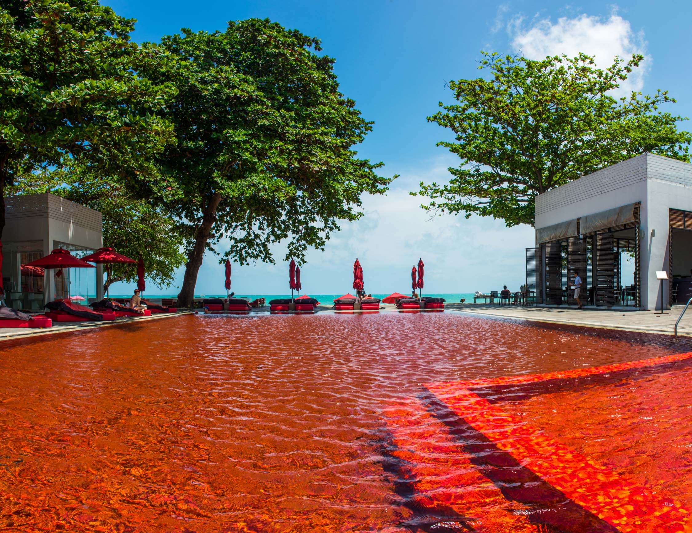 People relax on lounge chairs by a red-tiled pool under large green trees and a clear blue sky at a beach resort.