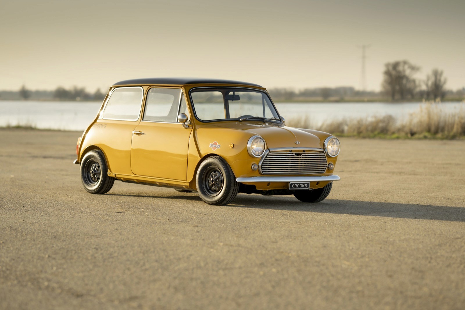 A vintage yellow mini classic car parked on an open road with a scenic background of water and trees.