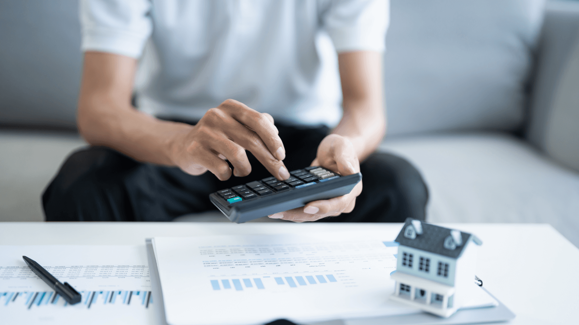 Eine Person sitzt auf einem Sofa und benutzt einen Taschenrechner, während sie von Papieren umgeben ist und ein Stift auf dem Tisch liegt.