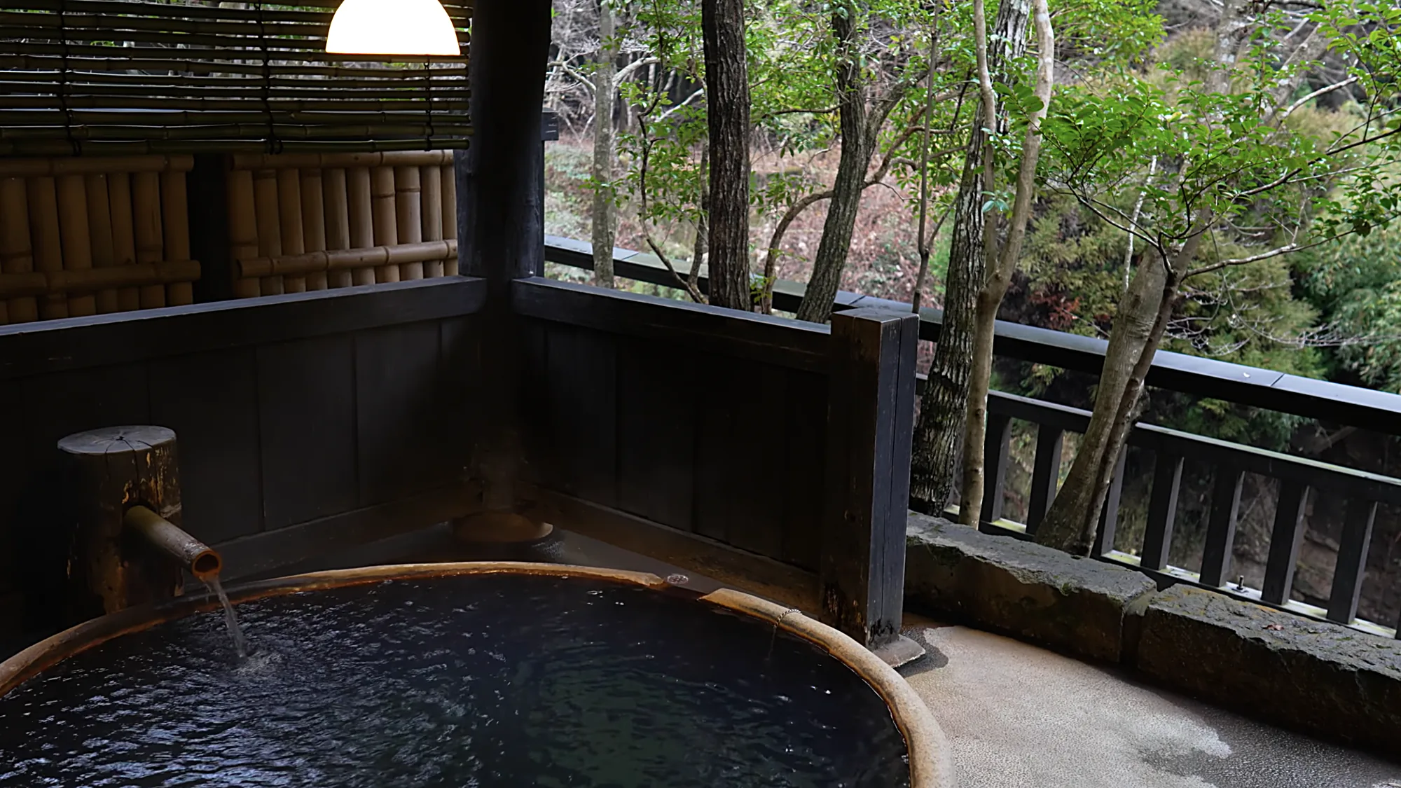 A round timber tub at Kurokawa Onsen with a bamboo spout, a timber rail and forest behind
