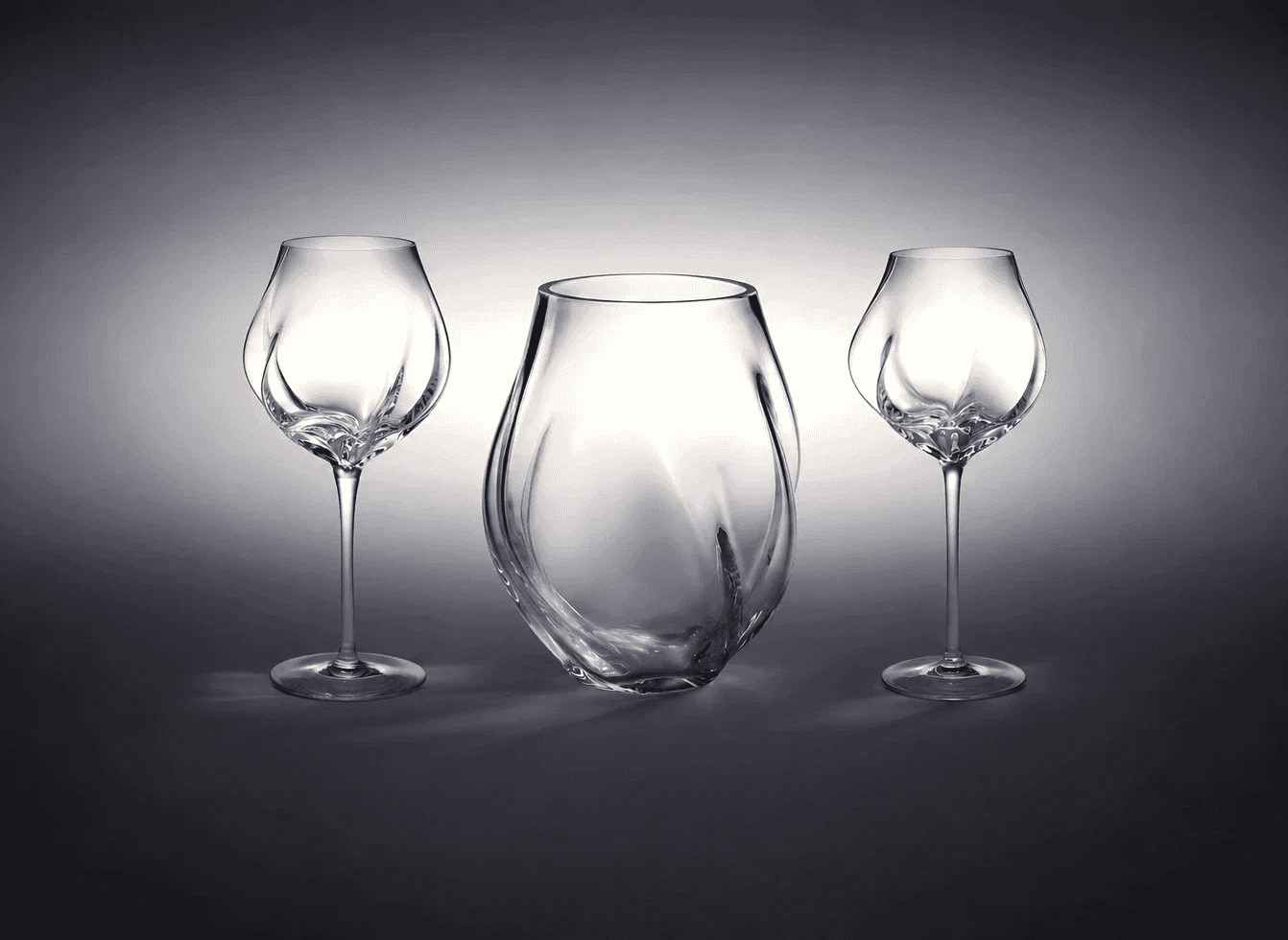 Two wine glasses flanking a clear glass chilling bucket, all three carrying the same undulating form, on a gray gradient.