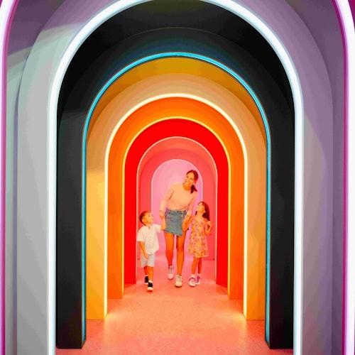 一位女士和兩個小孩走過一個充滿明亮色彩且燈光閃爍的拱門隧道。