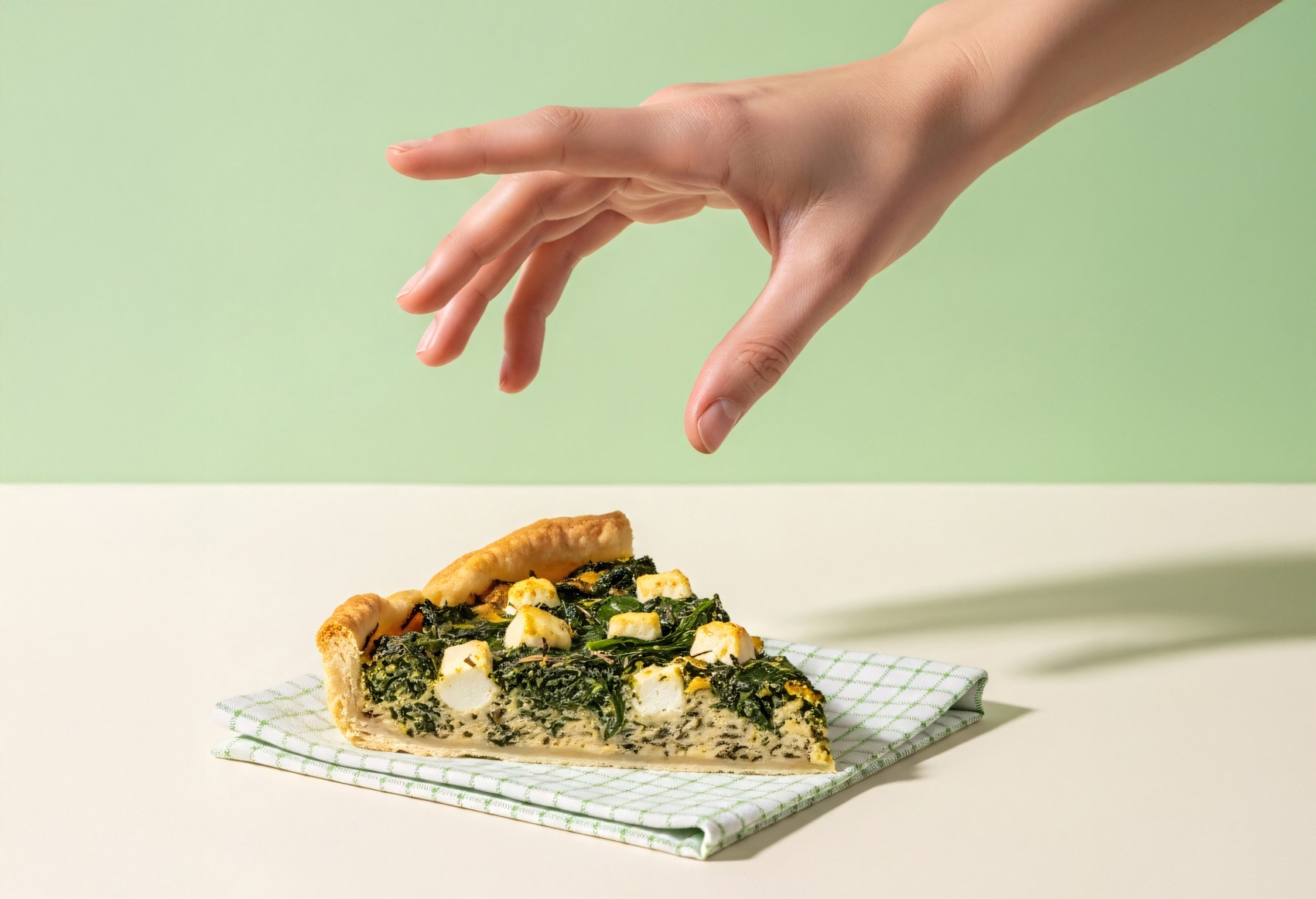 A hand reaches for a slice of spinach and feta quiche resting on a green checkered napkin atop a white table, set against a light green background.