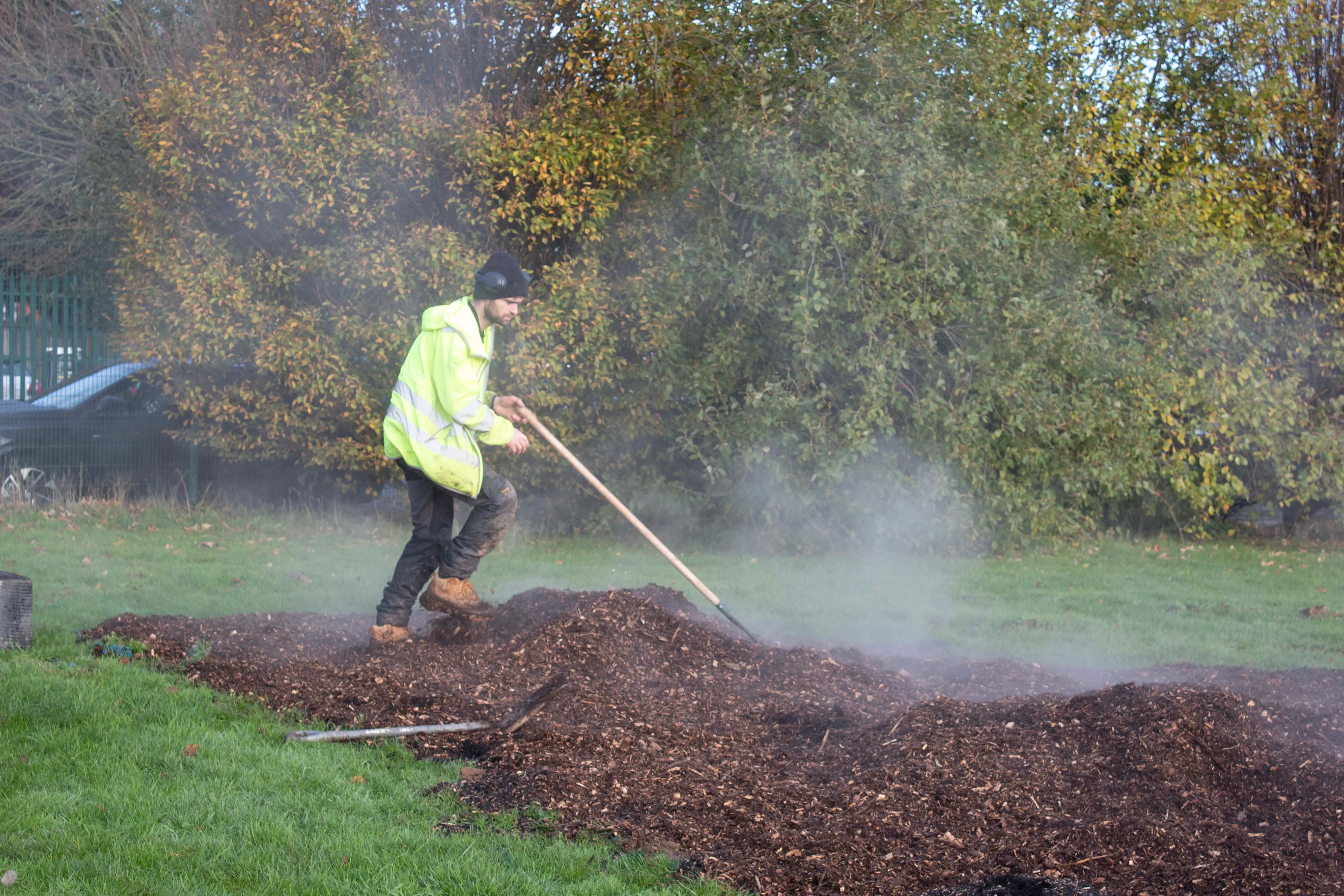 A person in a yellow jacket is using a shovel to spread mulch in a grassy area, with trees in the background.