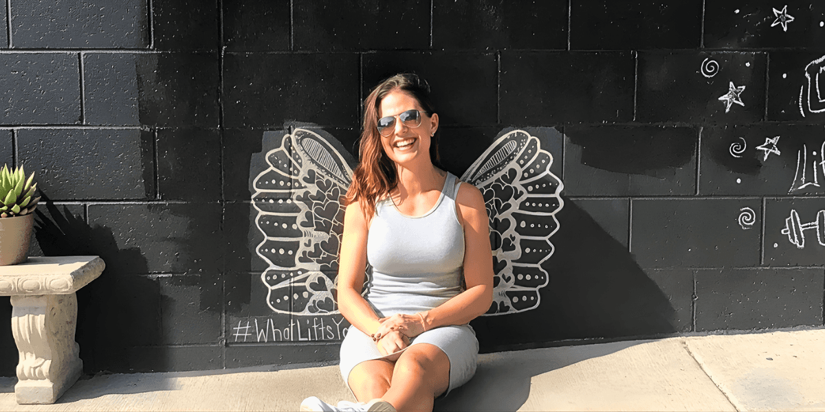 Angela Porrelli sitting in front of a mural with illustrated wings, smiling outdoors in sunglasses.
