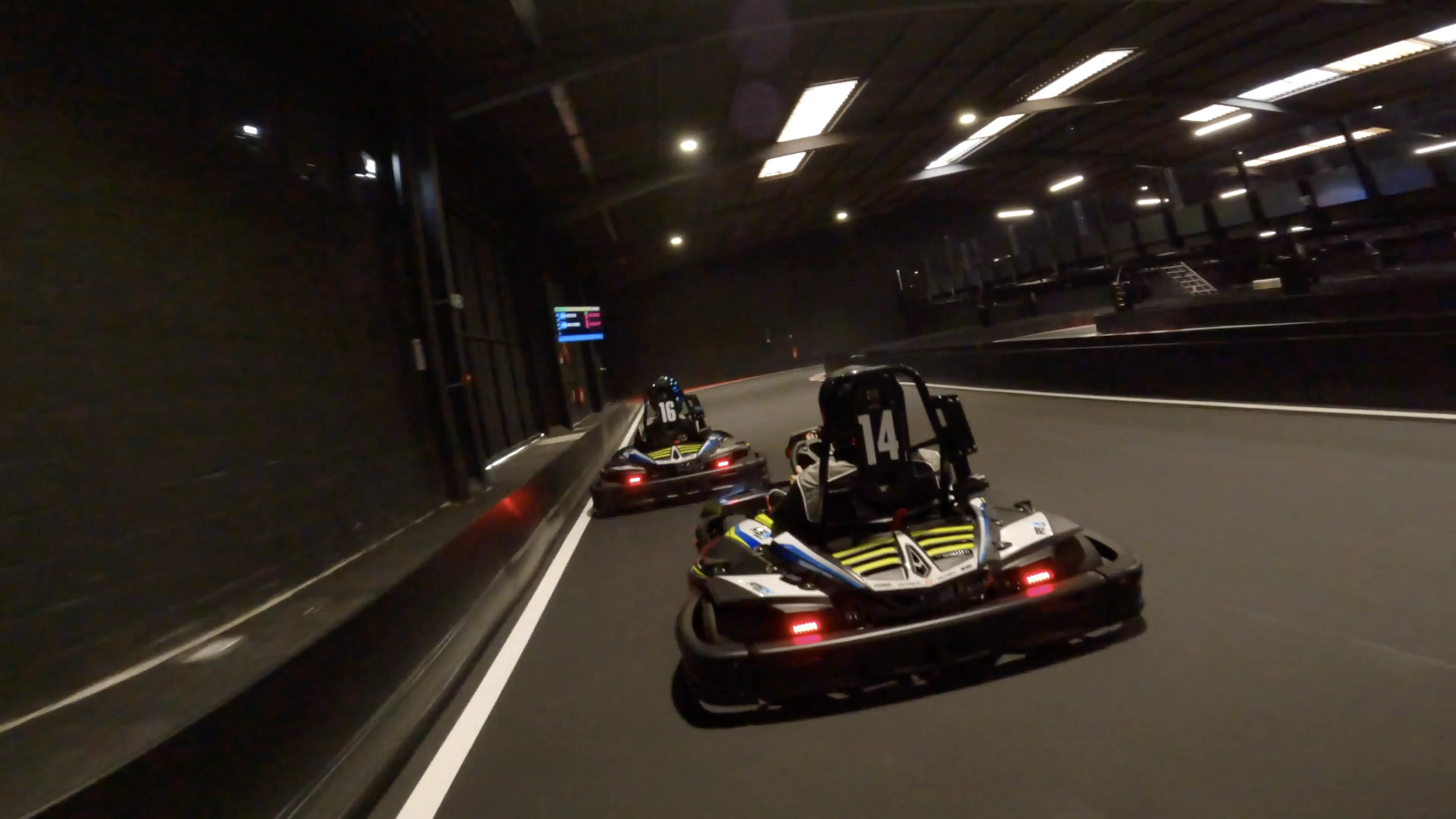 A pair of go-karts speed around an indoor track with dim lighting, showcasing dynamic racing action and capturing the thrill of go-karting excitement.
