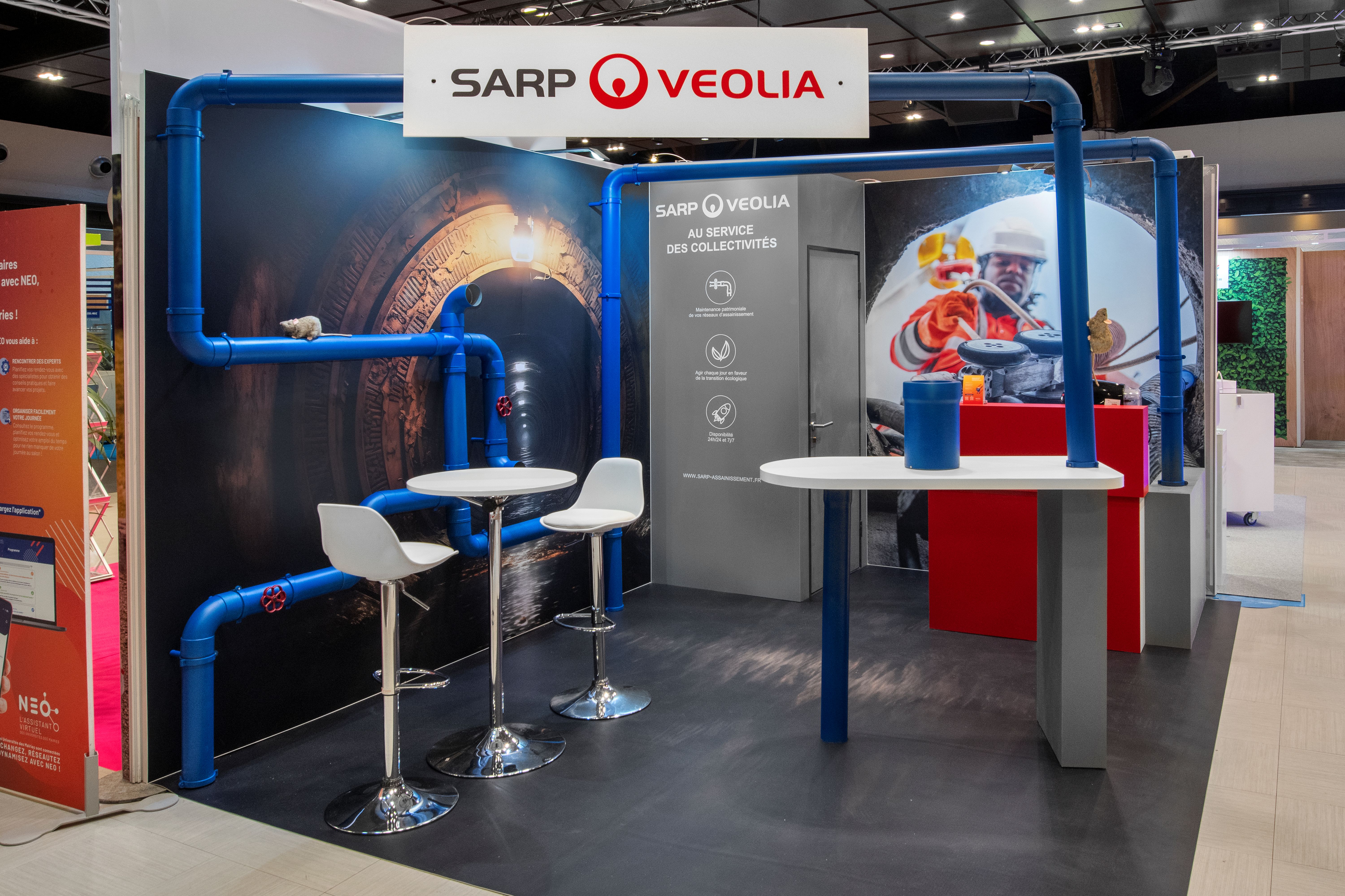 Stand SARP Veolia UMOP 2025 conçu par Expace – vue latérale du stand avec installation de tuyauteries bleues décoratives, podiums rouge et gris et visuel immersif réseau souterrain