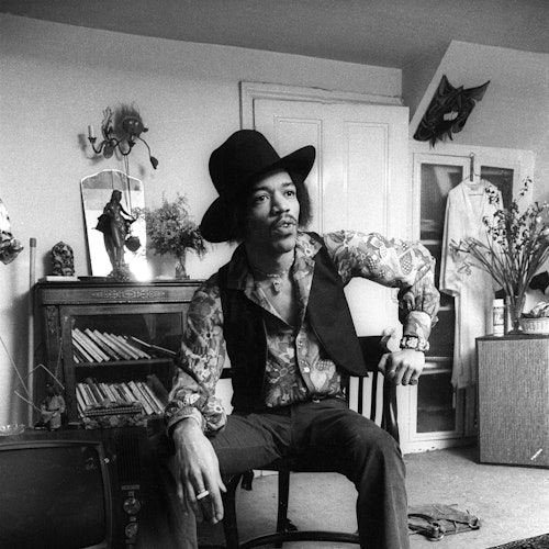 Un hombre con un sombrero y una camisa estampada se sienta en una silla en una habitación desordenada con un gabinete, libros y un televisor.