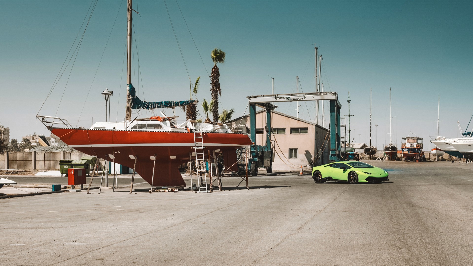 Green Lamborghini Huracán parked in a boatyard beside a red sailing yacht up on stands, with a travel-lift crane, workshop and masts of moored boats under a clear sky - a marina setting