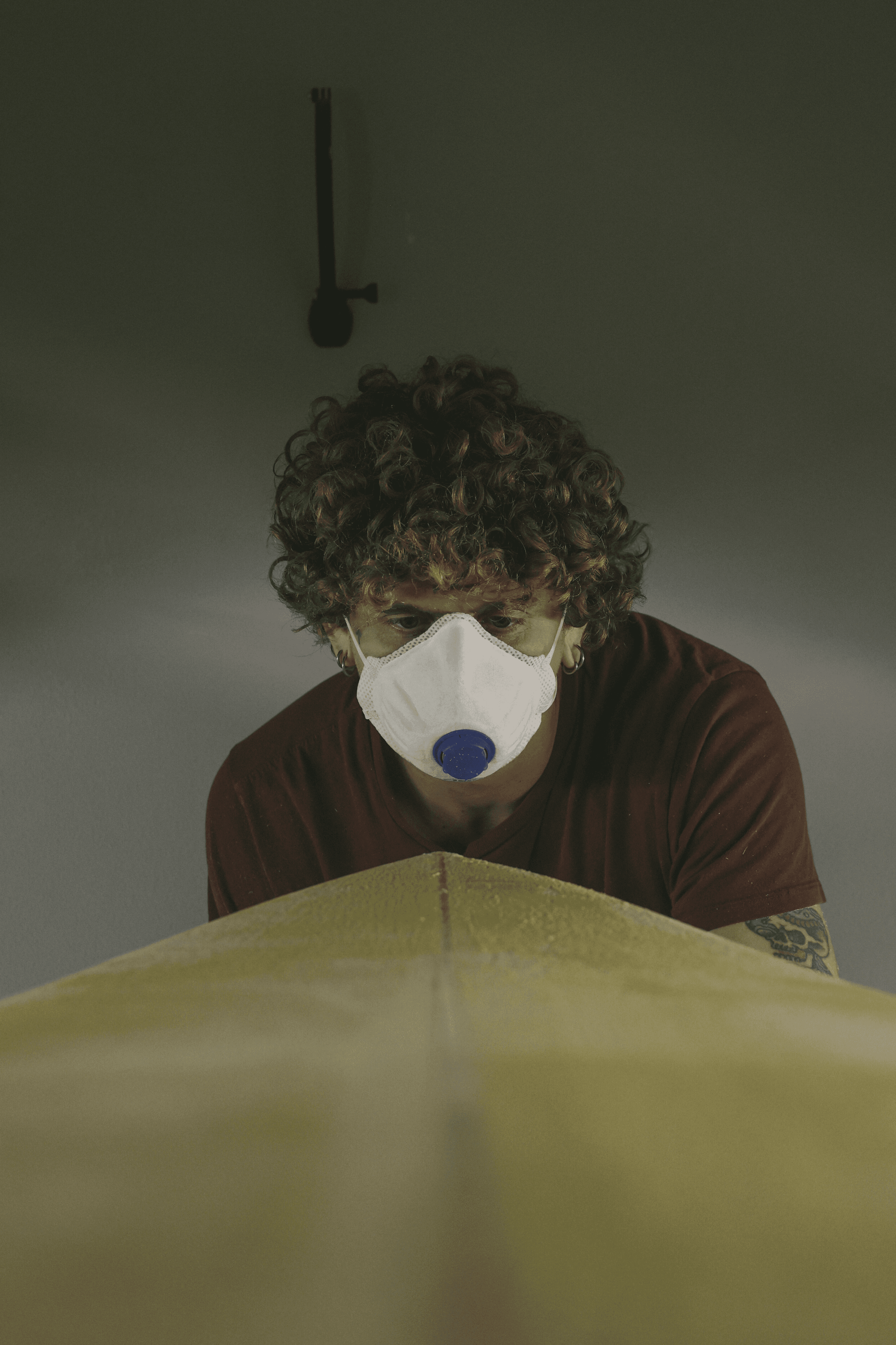 Giovane artigiano (shaper) con capelli ricci e mascherina, concentra lo sguardo sul bordo di una tavola da surf gialla.