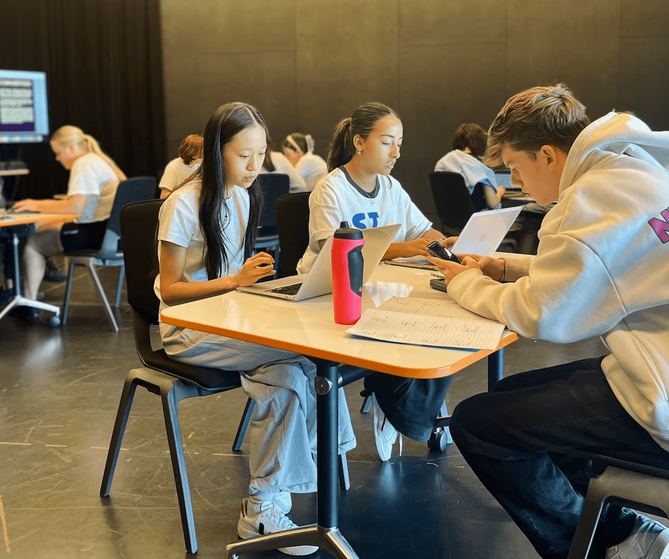 Groups of teenagers participate in a classroom-style theatre workshop, working on laptops and phones at tables in a studio space, with a presentation projected on a screen in the background.