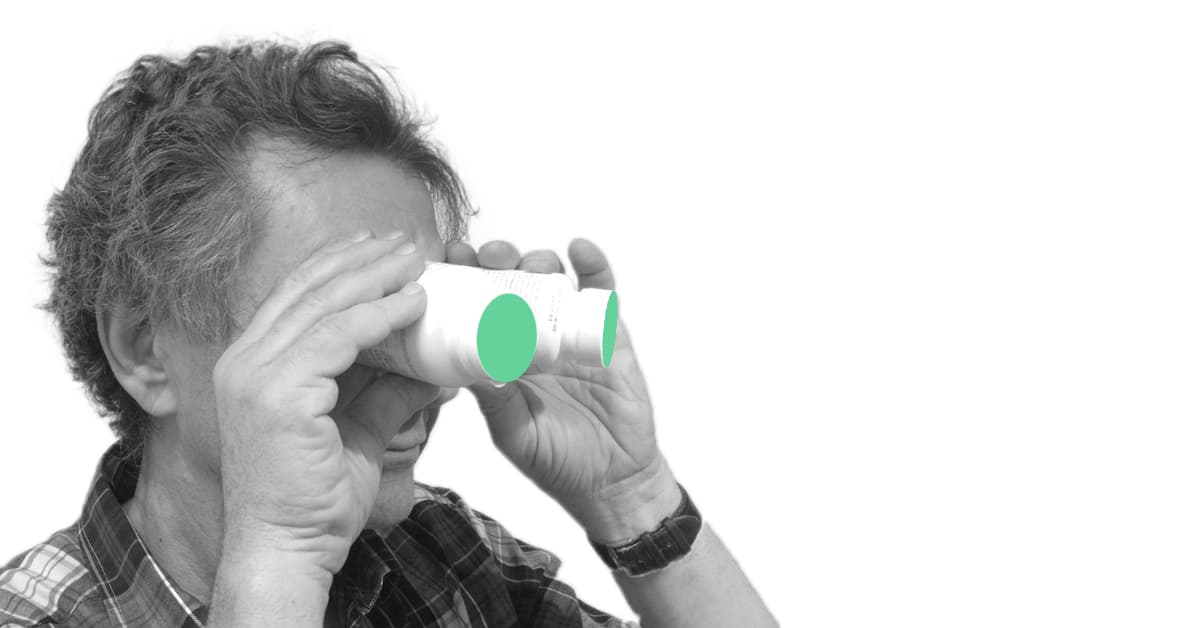 Older man with curly gray hair looking through two white medicine bottles held like binoculars; the bottle ends are marked with green circles. The image is black and white except for the green accents.
