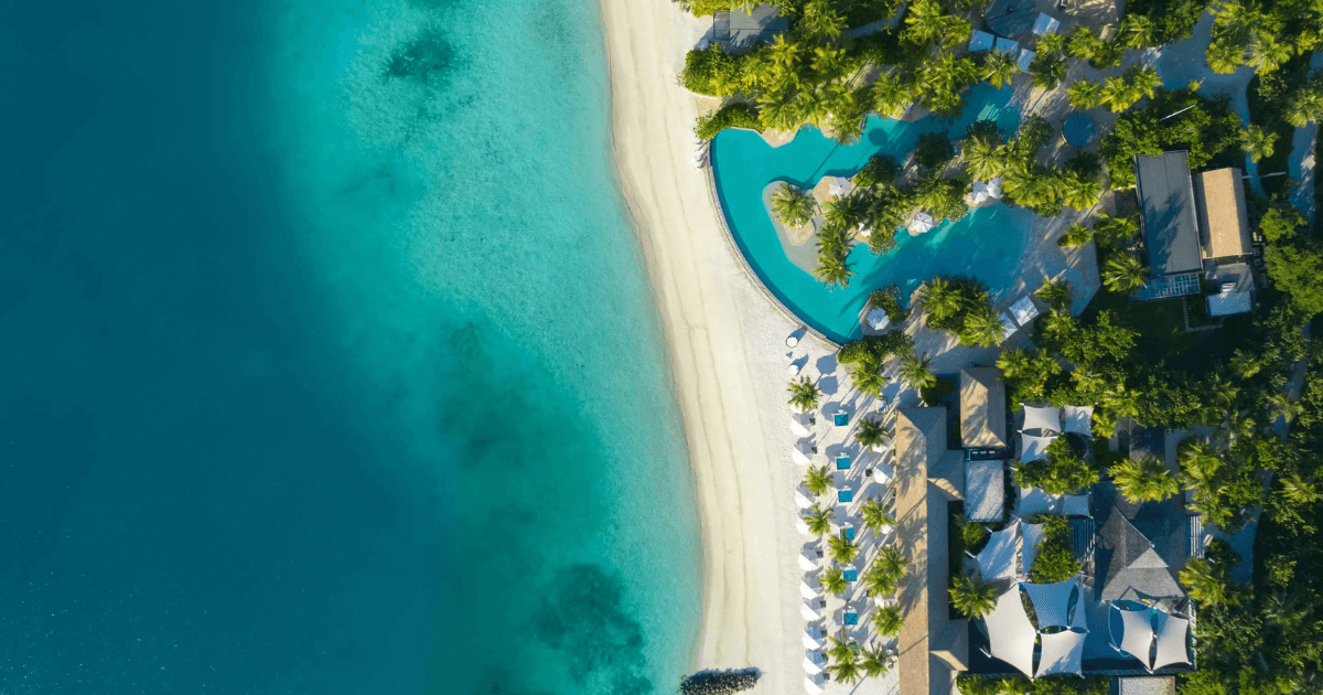 Aerial view of a beachfront resort with a pool, sun loungers, and turquoise ocean water along a sandy shore.