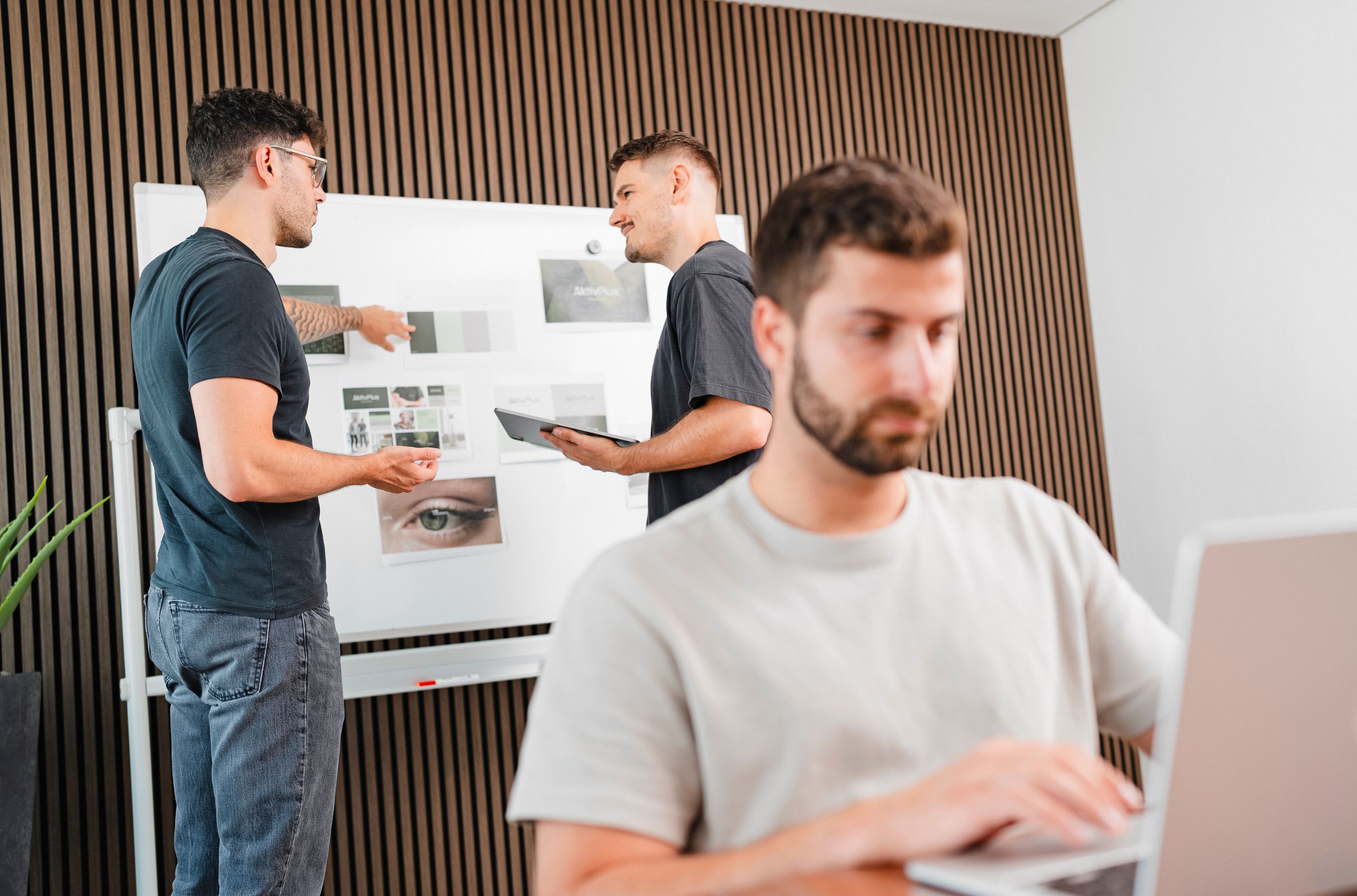 Jannis Schuler präsentiert Design-Referenzen am Whiteboard, Kollege arbeitet im Vordergrund am Laptop