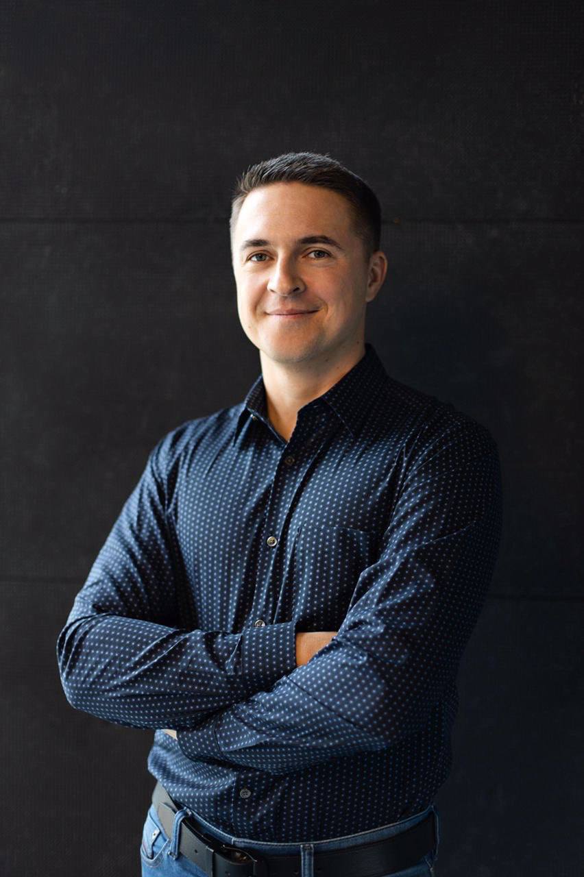 Alex, Level Events (custom trade show booth builders) co-founder in a dark, dotted shirt smiling with arms crossed against a plain, dark background.