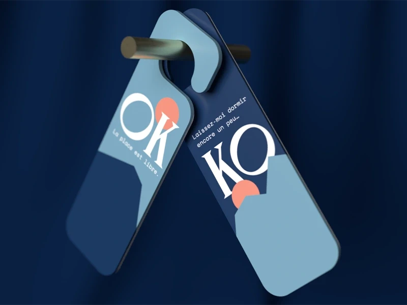 Accroche-portes de l’Hôtel Outa au design graphique bleu, avec les messages typographiques “OK, la place est libre” et “KO, laissez-moi dormir encore un peu”, suspendus à une poignée.