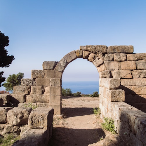 Des ruines d'une arche en pierre avec un chemin de terre menant à une vue sur la mer sous un ciel bleu clair. Des arbres et des buissons sont visibles.