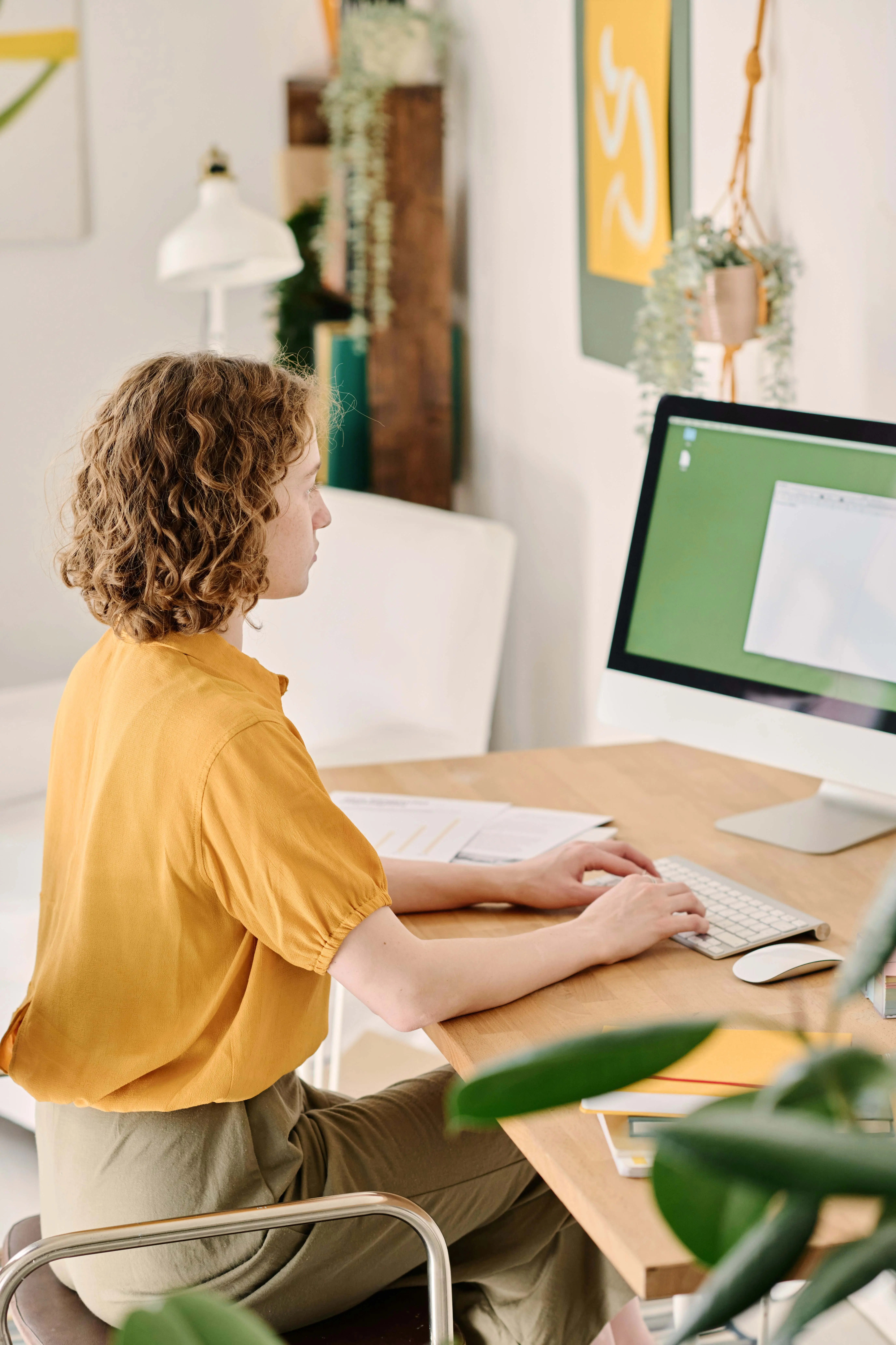 Person with curly hair wearing a yellow blouse sitting at a wooden desk, typing on a keyboard in front of a desktop monitor displaying a document.