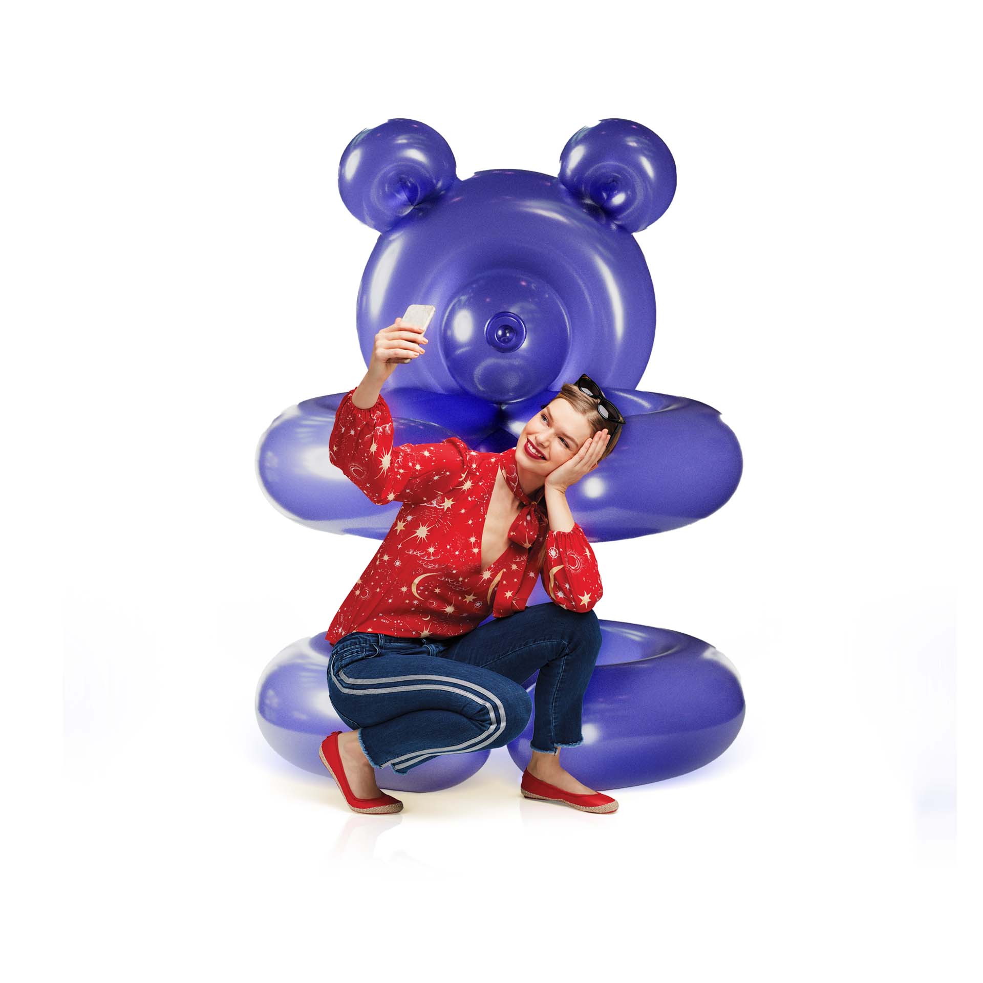 A woman happily sits on a glossy, oversized balloon sculpture of a teddy bear in a rich coral shade, showcasing a pair of Butterfly Twists ballet flats in a colorful pattern.