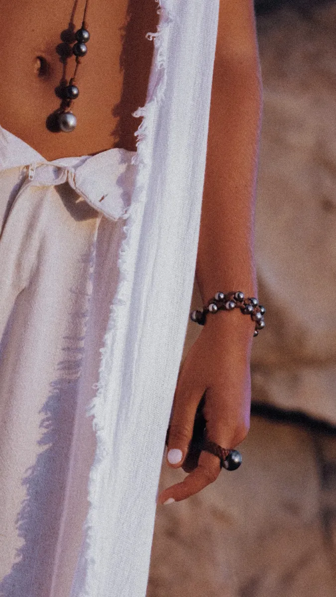 Close-up of a woman in a frayed white dress wearing a long Tahitian pearl necklace and a multi-strand pearl bracelet wrapped around her wrist, paired with a bold pearl ring — wear a piece of the ocean, born in Saint Barth. Handmade luxury jewelry boutique — Manos de Oro Saint Barth.