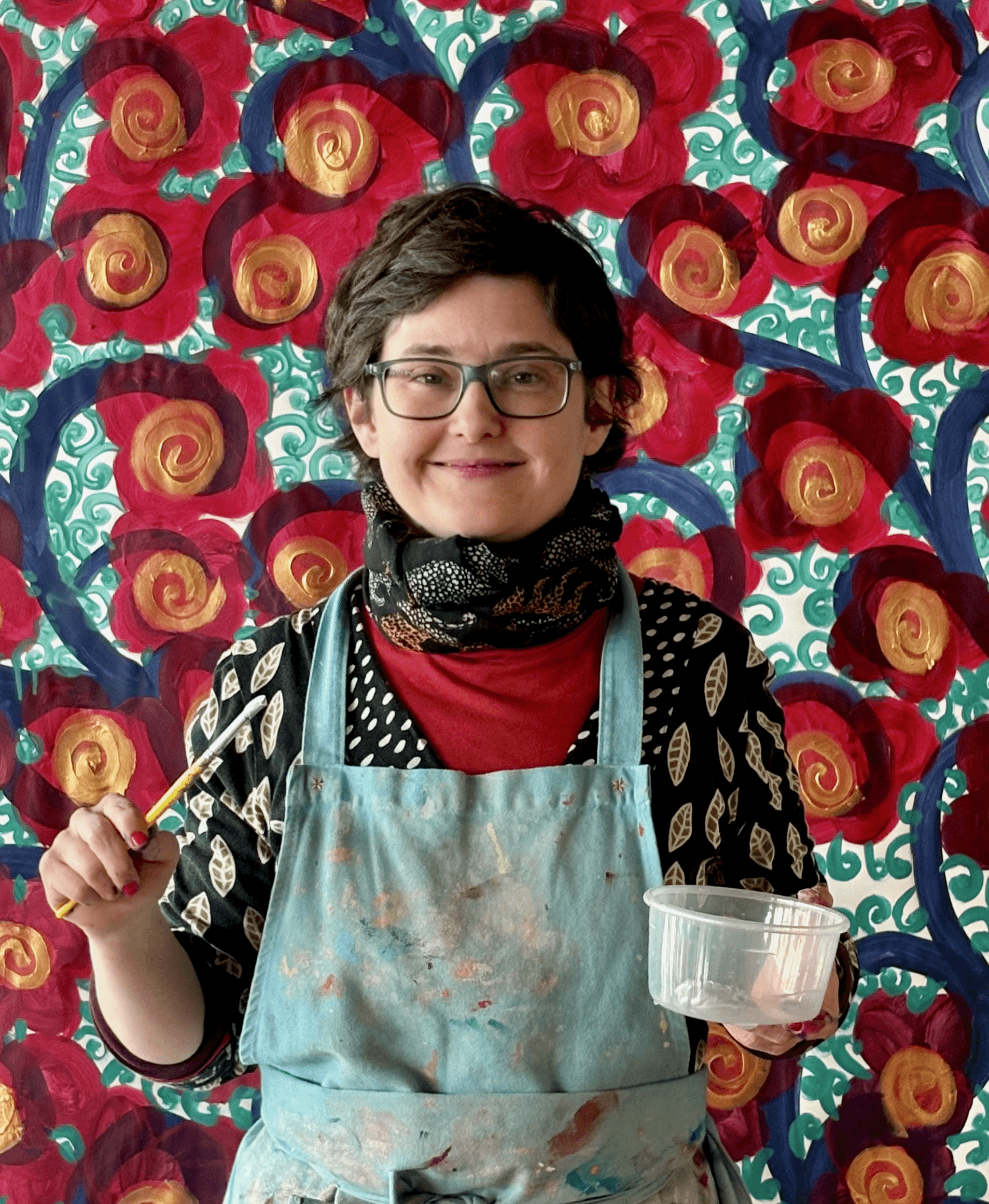 Portrait de Morgane Salmon, souriante et vêtue d’un tablier taché de peinture, tenant un pinceau et un petit récipient devant un décor peint de roses rouges aux spirales dorées et feuillages turquoise.