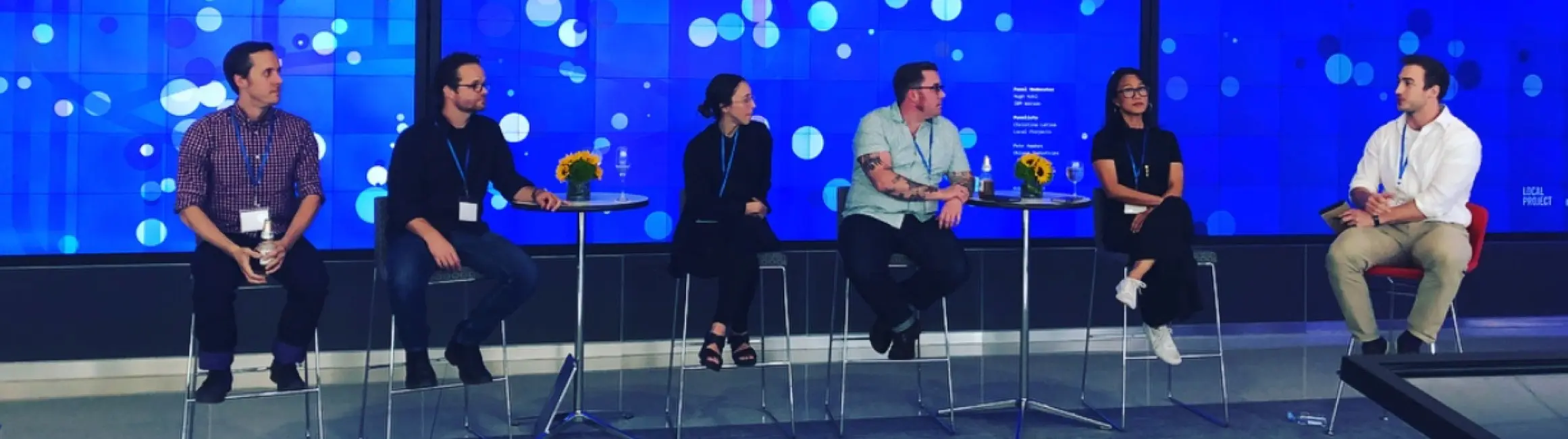 Six panelists seated on high stools in front of the Watson Experience Center's immersive wall displaying blue generative visuals. From left to right: Fred Benson, Pete Hawkes from Oblong Technologies, Christina Latina from Local Projects, Rob Harrigan, Jenny Woo from IBM Watson, and Hugh Kohl — discussing how the team creates spatial AI experiences for client visits.