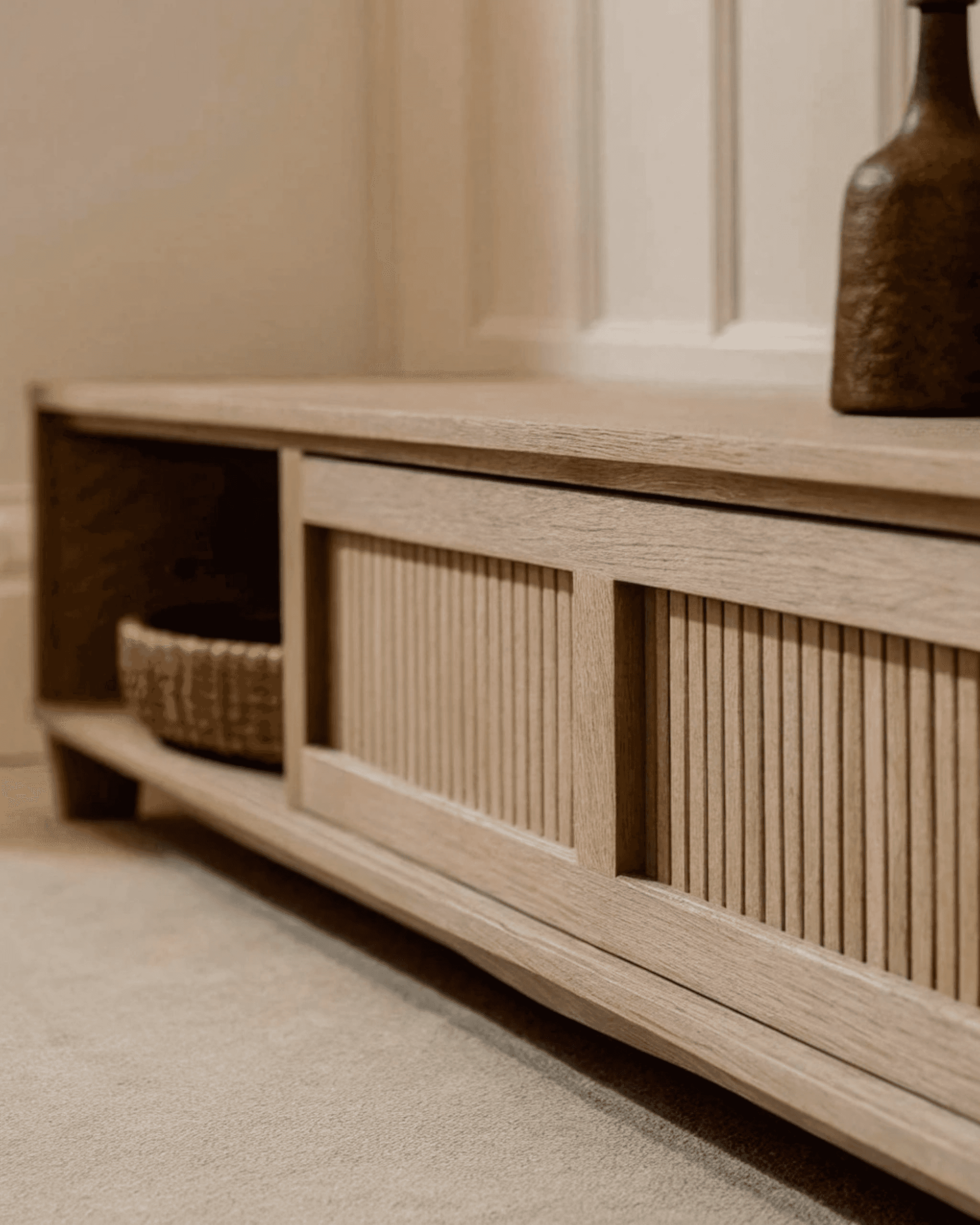 Modern wooden cabinet with slatted sliding door, woven basket, and ceramic vase on top, set against a white paneled wall.