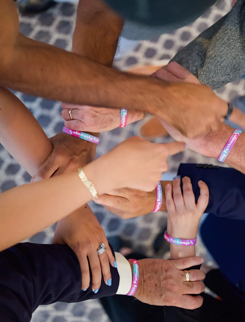 Hands joined together in a circle, each wearing a bracelet with the text “Fertility Pathways.”