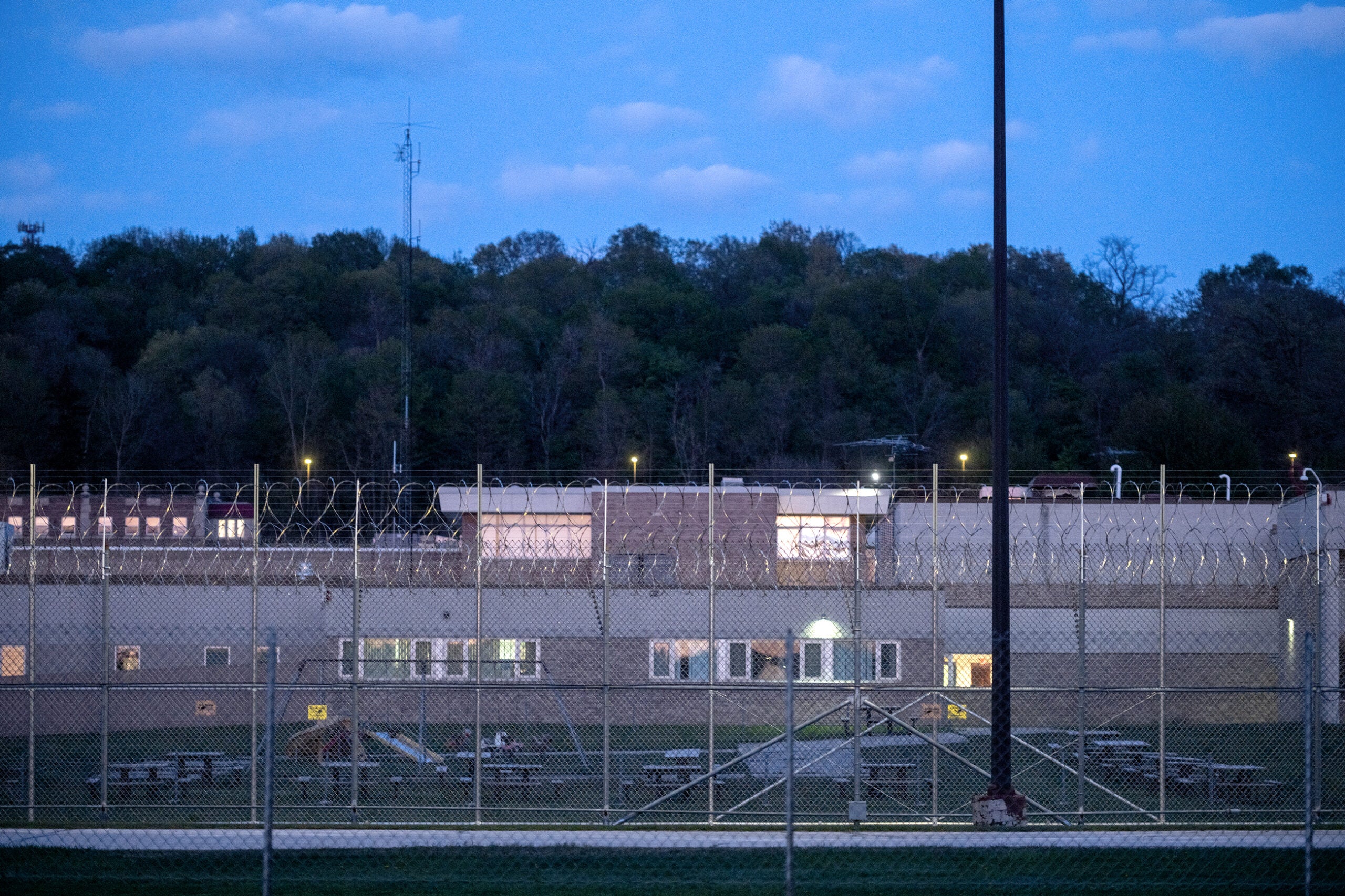 A fenced prison yard with barbed wire, picnic tables, and a building in the background, photographed at dusk with trees and a hill beyond.