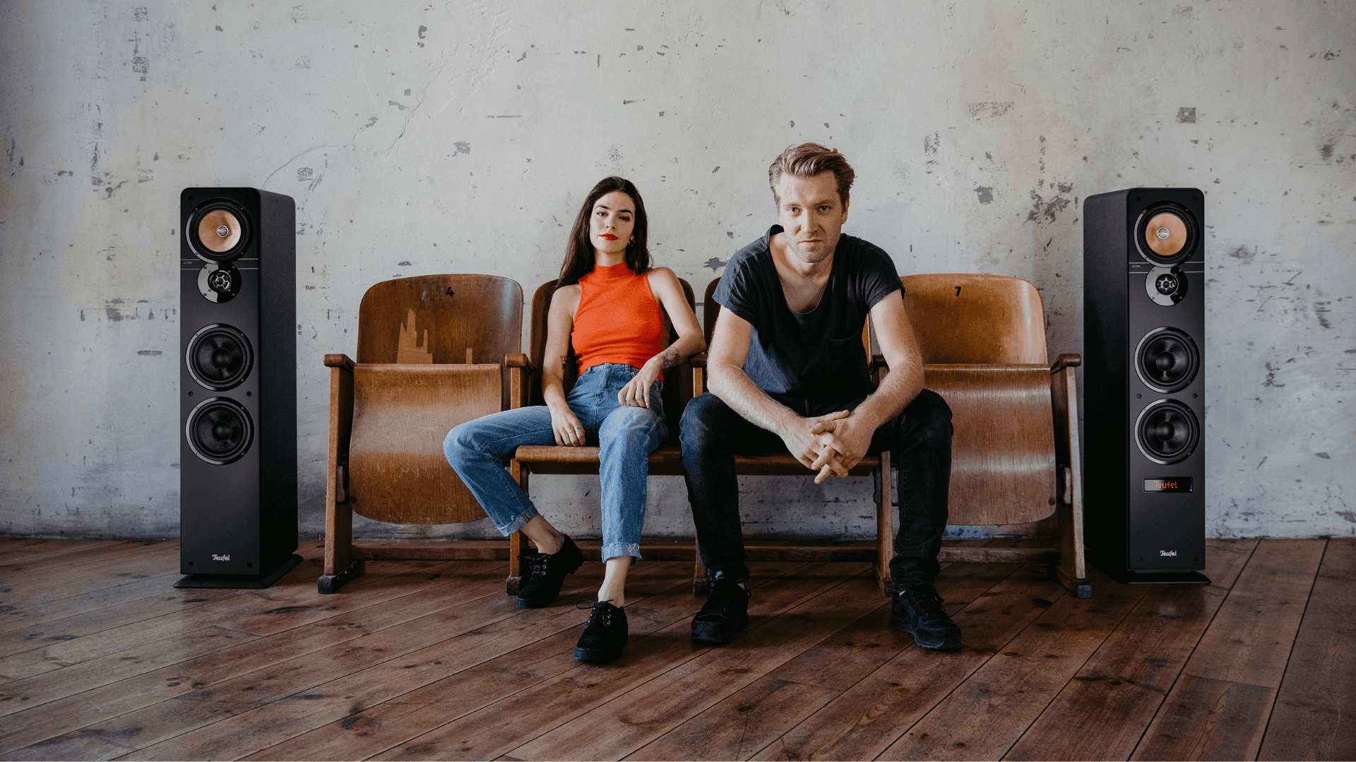 Two people sitting on wooden folding chairs between two Teufel speakers