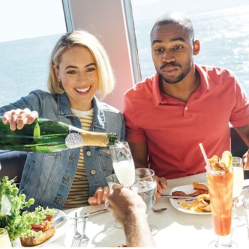 一名女士在餐桌上为三位顾客倒香槟,背景是一望无际的海景。