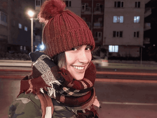 A smiling person in a red beanie and scarf stands on a city street at night. Buildings are visible in the background.
