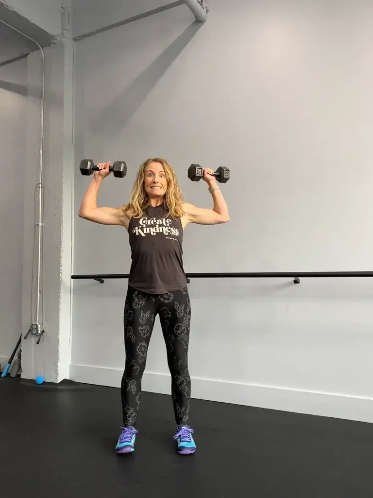 Heather Hausman with long hair is lifting dumbbells in a gym, wearing a tank top that says "Create Kindness," paired with patterned leggings and blue sneakers, against a minimalist background with a bar and exercise equipment.