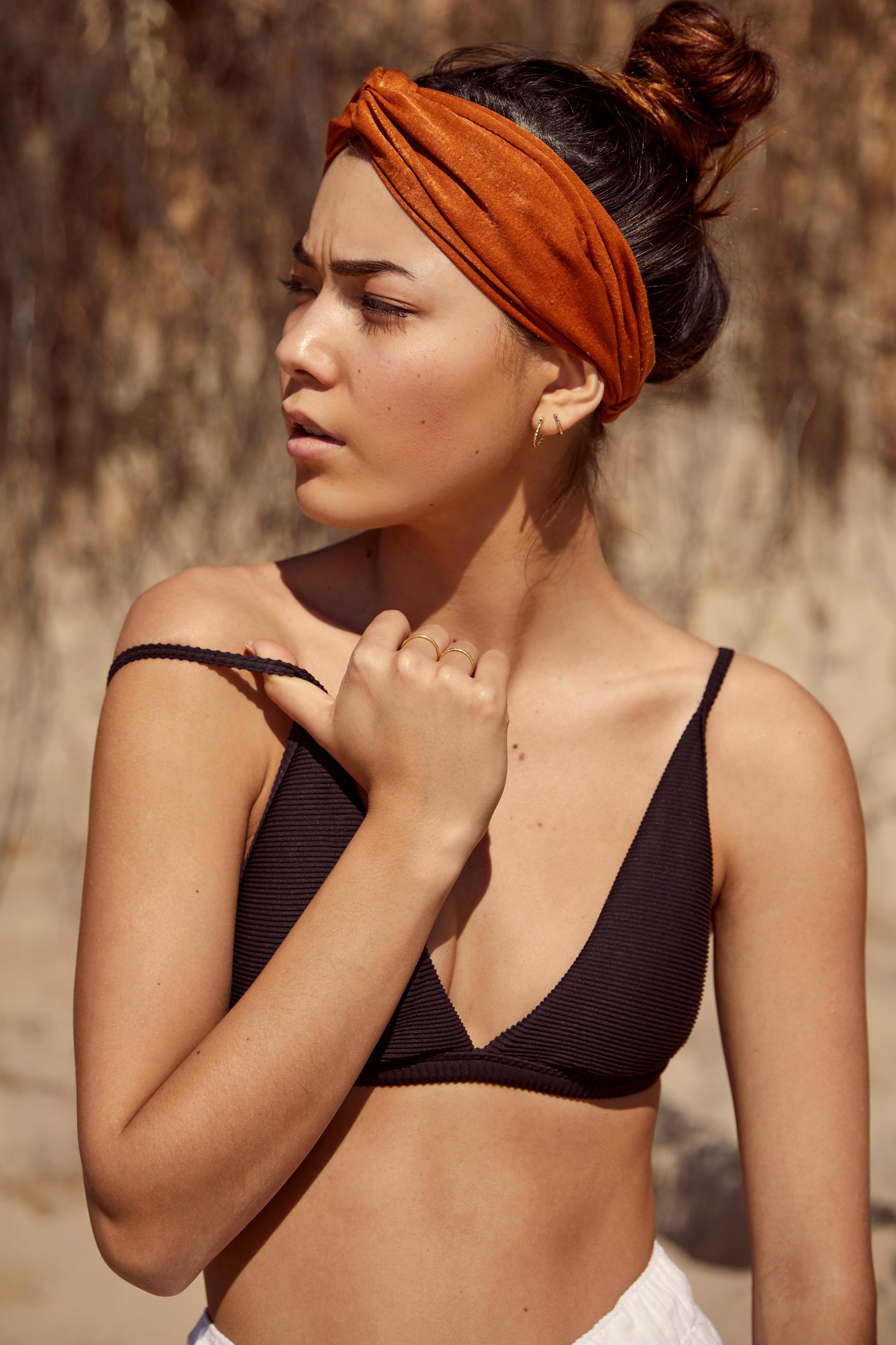 A close-up fashion photo showing a female model pulling at the shoulder strap of her black bikini top while squinting in the bright light and heat.