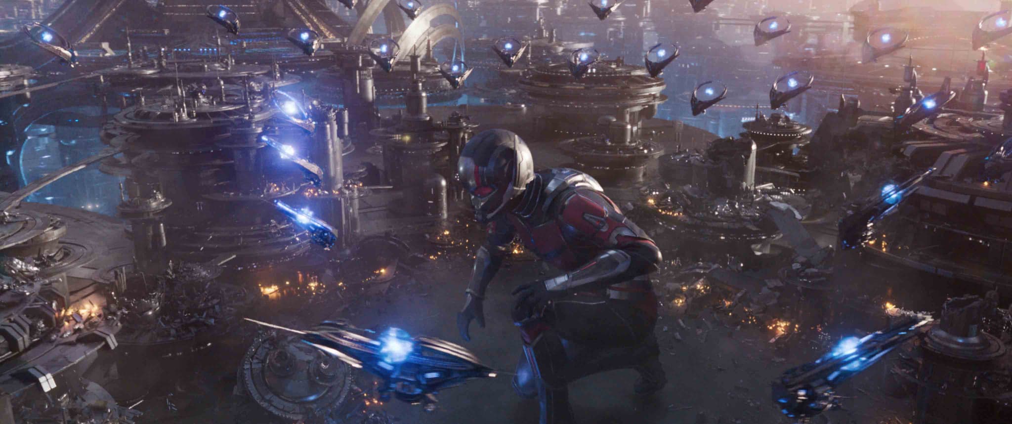 A giant person in a futuristic suit stands among a sprawling sci-fi cityscape under attack. Flying ships with blue lights are visible, and explosions illuminate the scene, creating a sense of high-tech warfare.