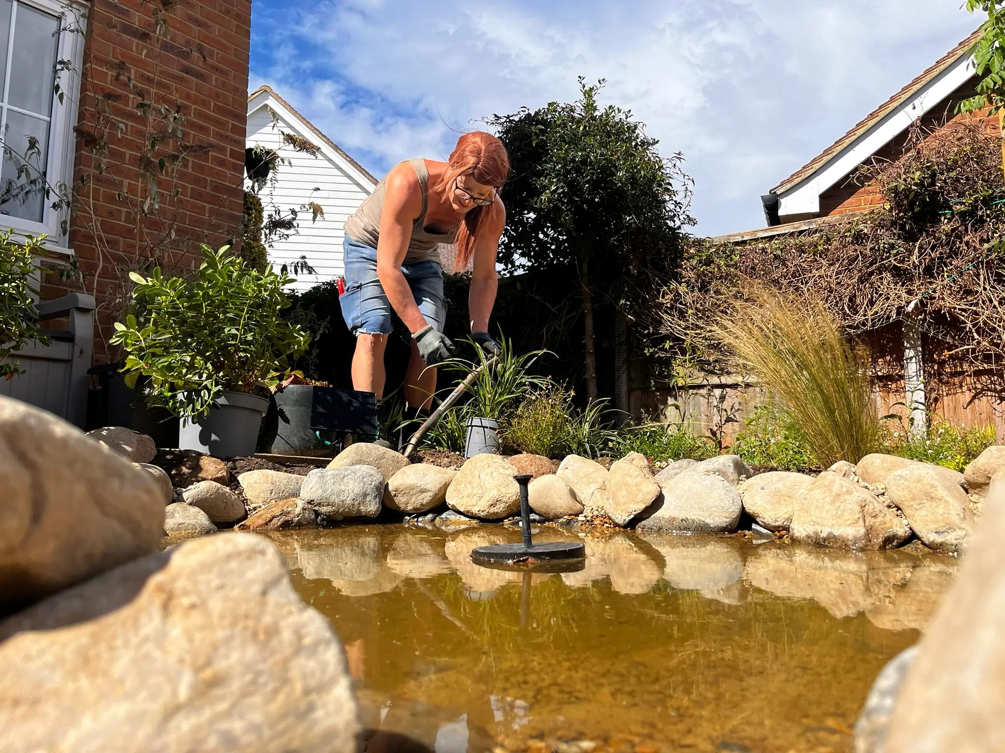 A person leans over a pond surrounded by rocks and plants, under a clear blue sky in a garden setting.
