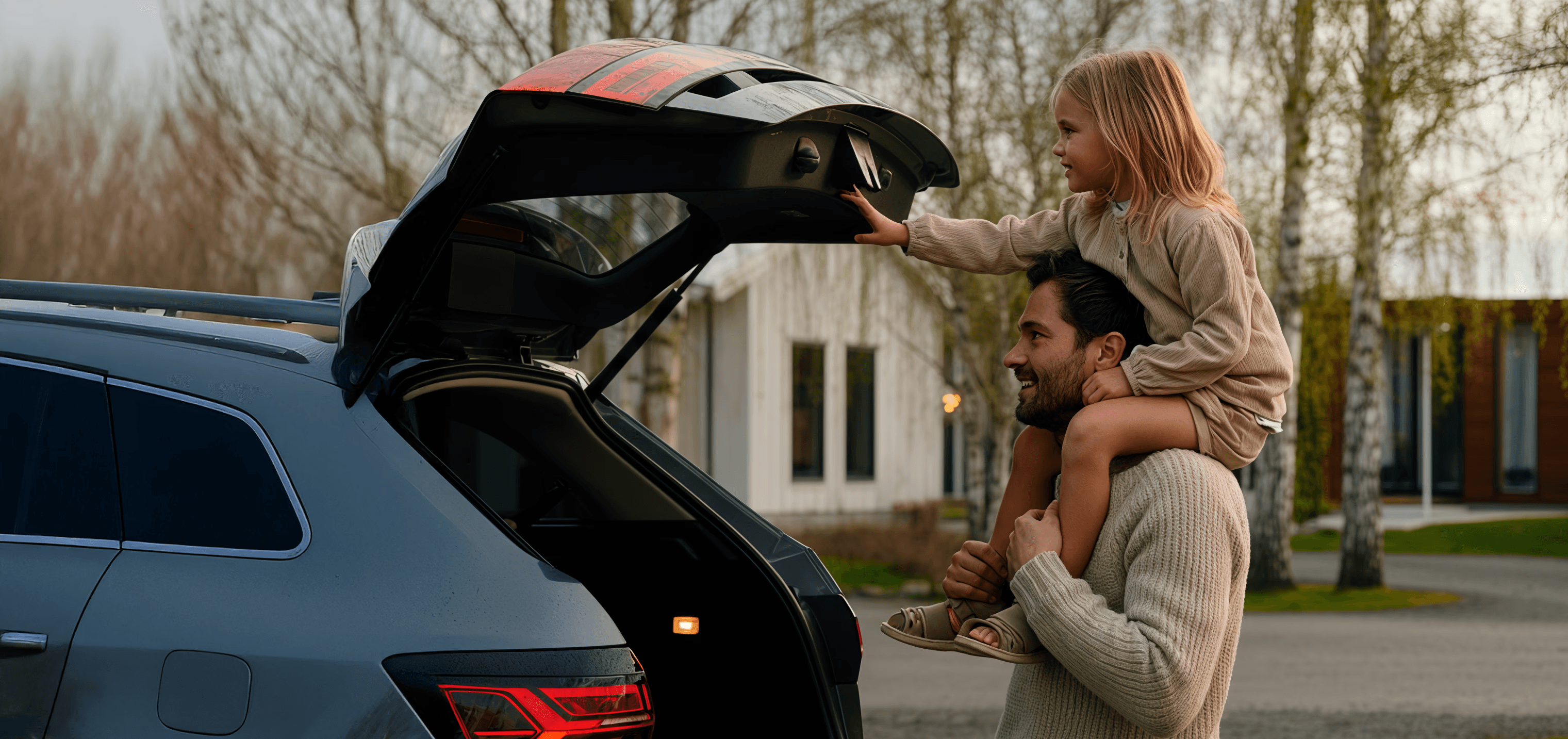 A father carries his daughter on his shoulders as she reaches into an open car trunk. They're by a car and some houses.