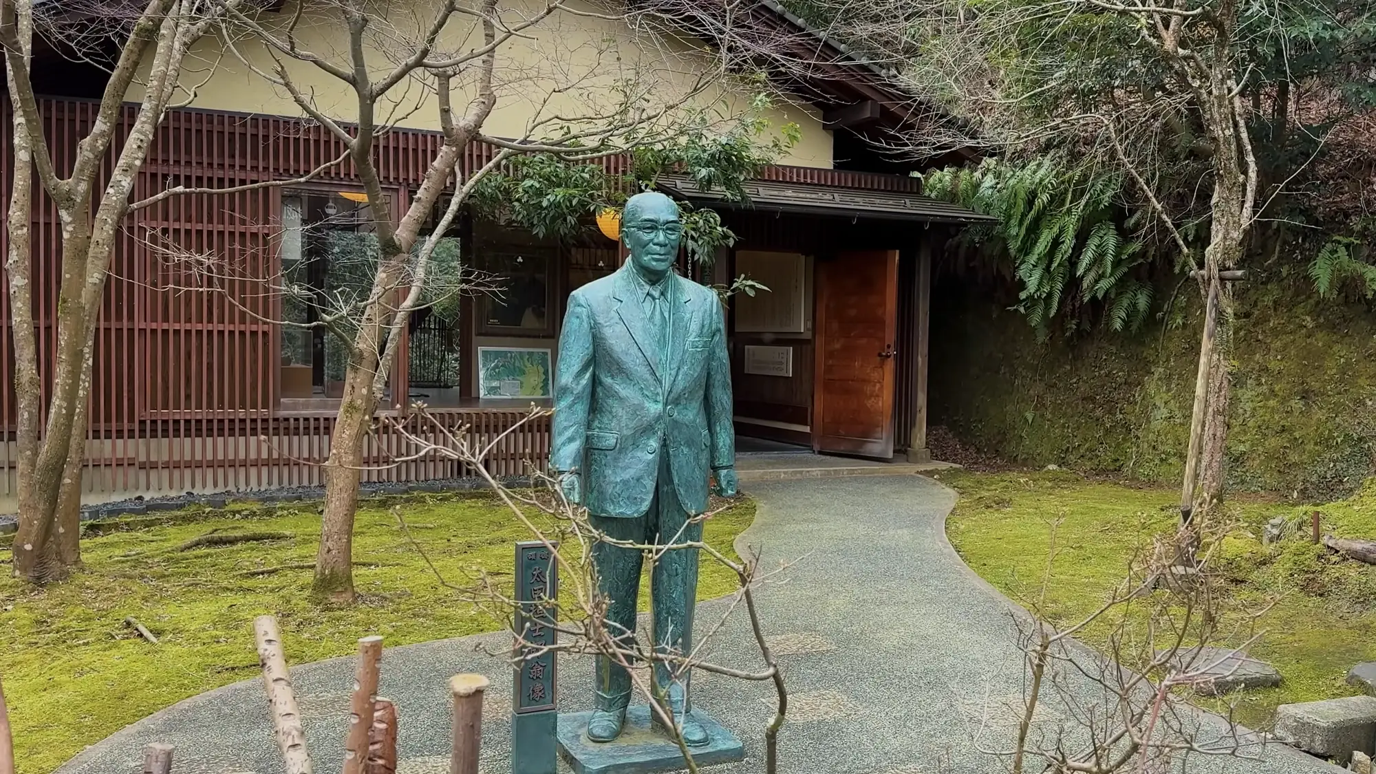 A bronze statue of a seated figure in a garden beside a timber building in Kinosaki.