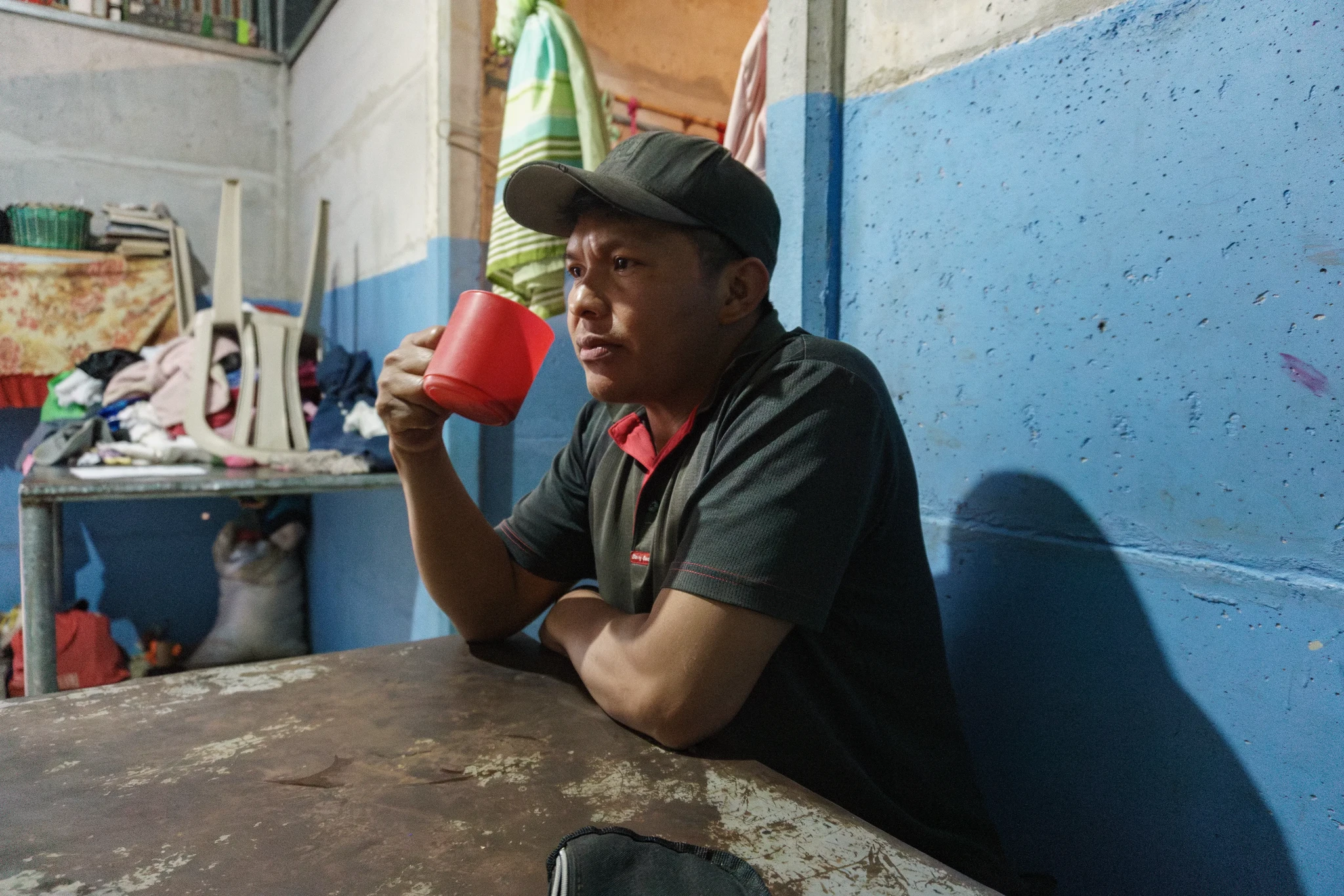 A man sits on the floor holding a toddler. The man appears to examine several identification cards in his hand. The child is shirtless, wearing red shorts, and looks off to the side. The room has a hammock and a rough concrete floor.