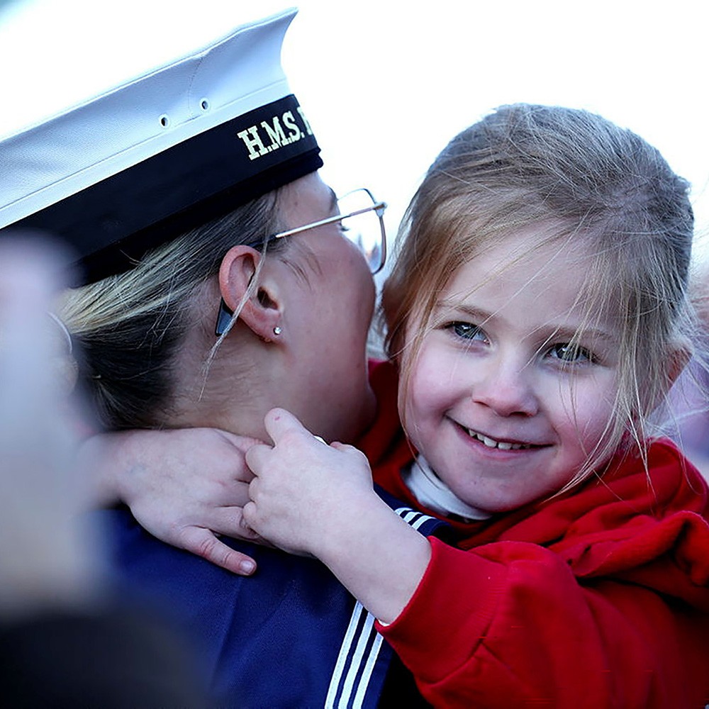 A young girl wearing a red jacket smiles at the camera while embracing a naval service member wearing an HMS cap and uniform. The service member, shown from behind, leans in close to the child in what appears to be an emotional reunion or tender moment, highlighting the personal connections and family bonds of military personnel.