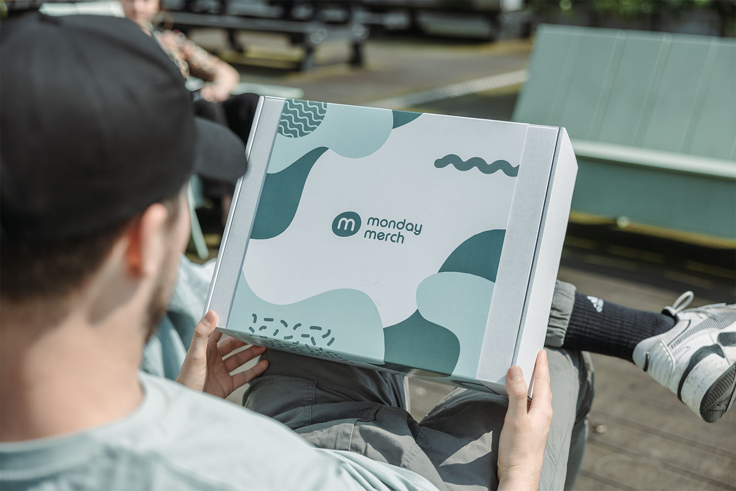 Personne tenant une boîte blanche avec des designs abstraits verts et le logo "Monday Merch"