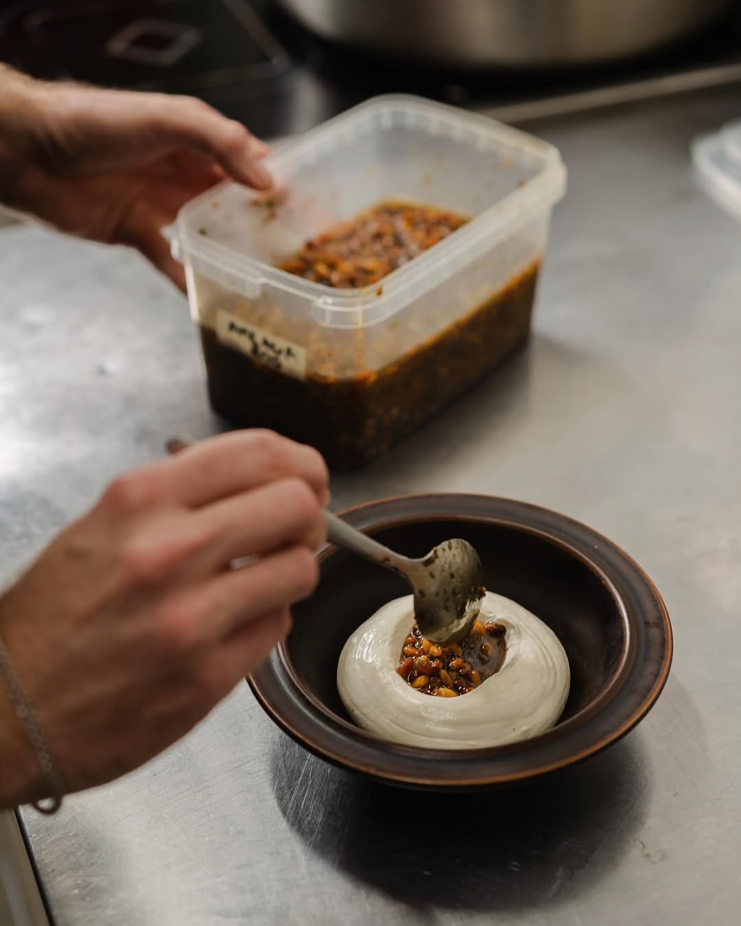 A group of five people dressed in casual, neutral-toned clothing sit around a rustic wooden table in a cozy, stone-walled kitchen, smiling and engaging in conversation with bowls and a covered dough resting in front of them, creating a warm, communal atmosphere.