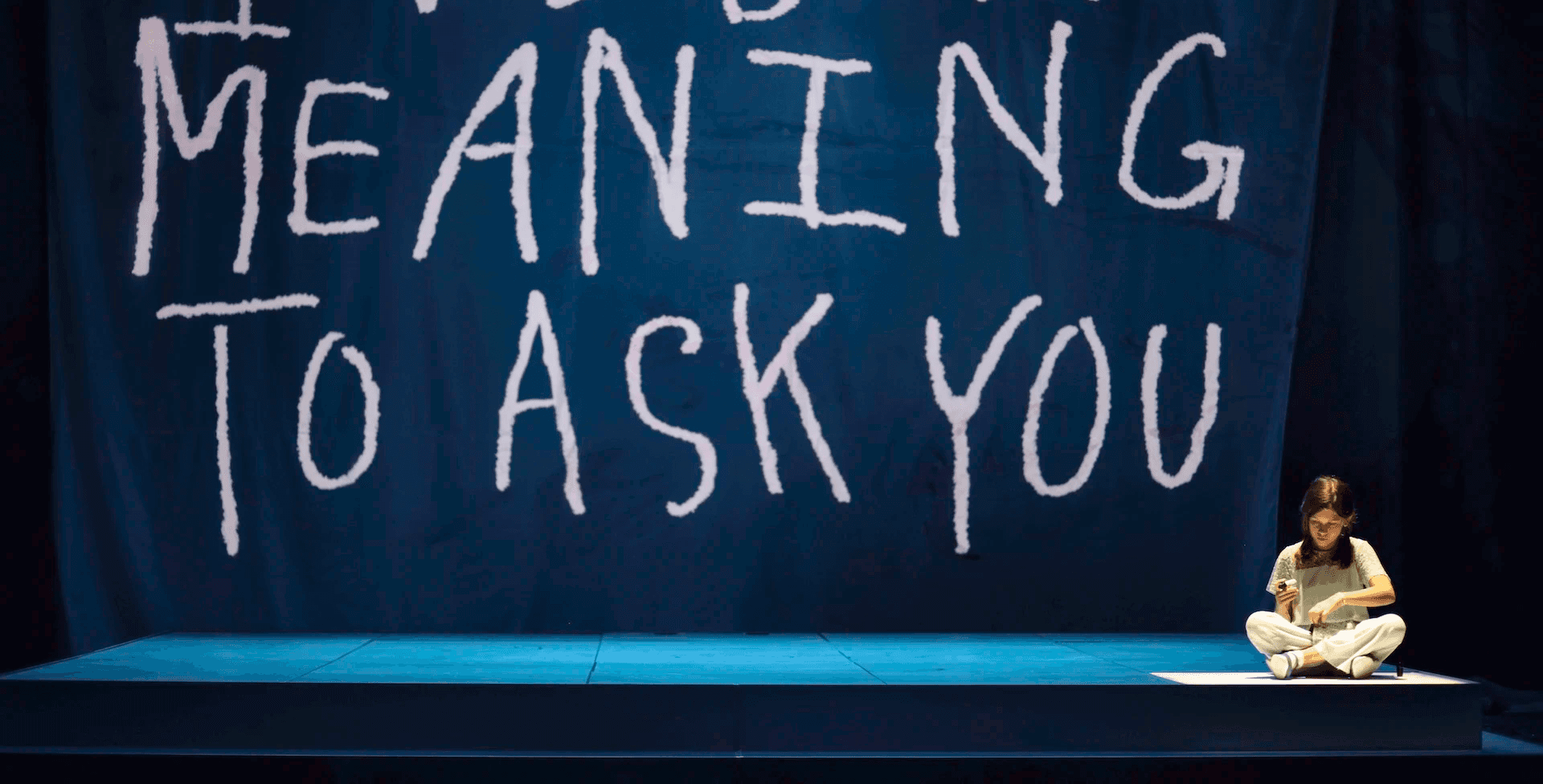A young performer sits cross-legged onstage holding a microphone, beneath a giant projected backdrop reading “I’VE BEEN MEANING TO ASK YOU.” The stark text and empty space around them highlight an intimate, reflective moment.
