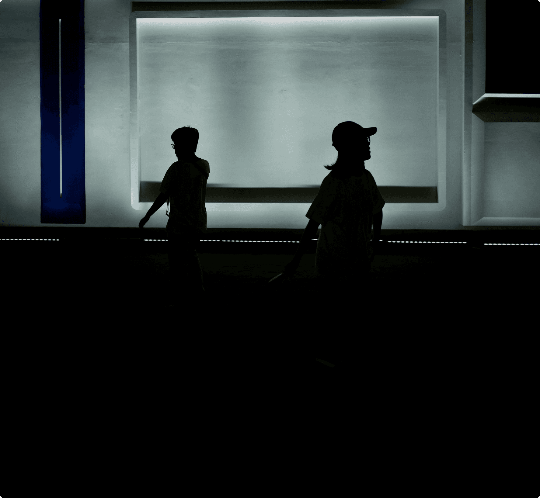 A silhouette of two people walking past a large, brightly glowing rectangular light fixture on a dark wall.