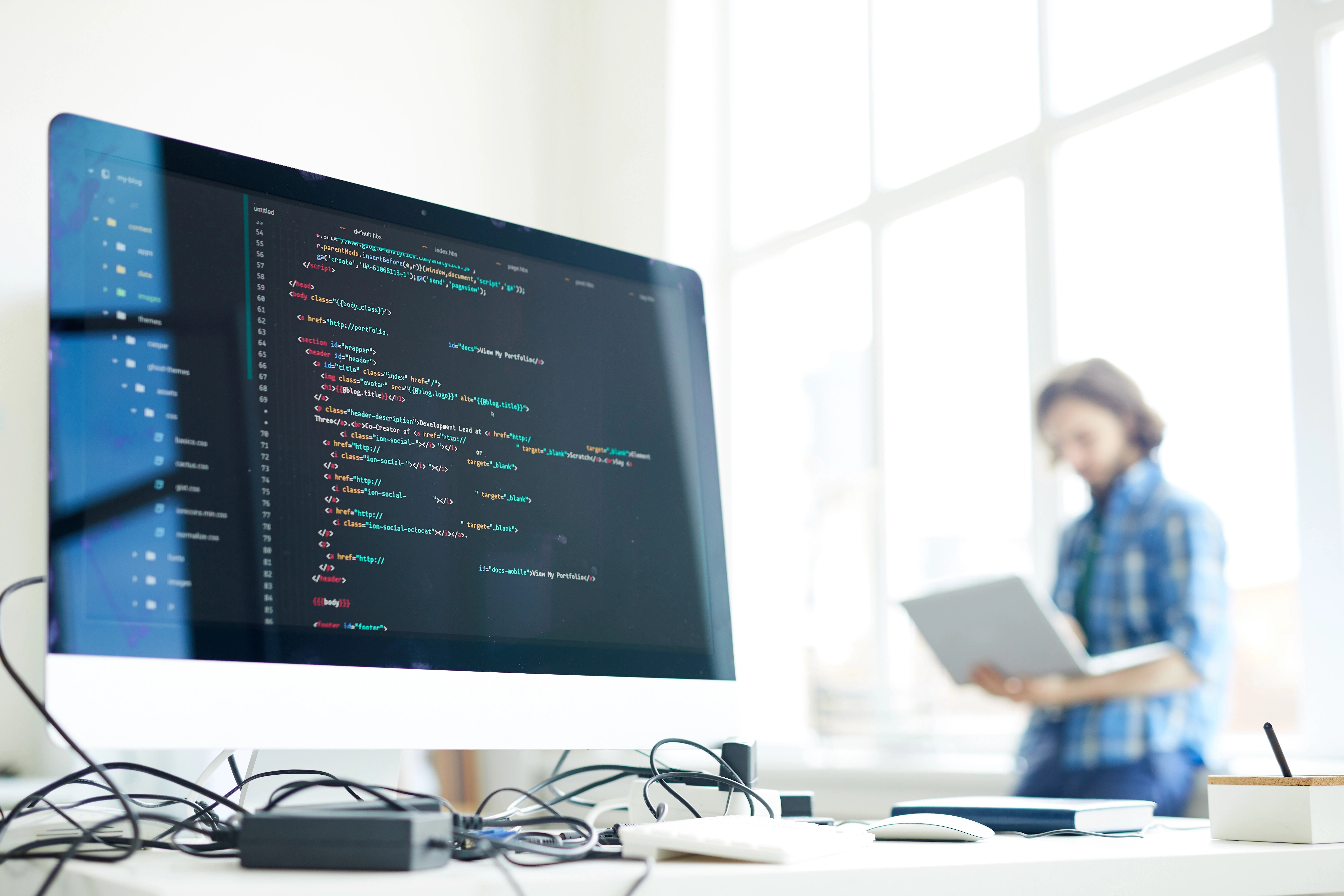 Developer working with a laptop in a bright office, with a desktop monitor displaying colorful code in the foreground